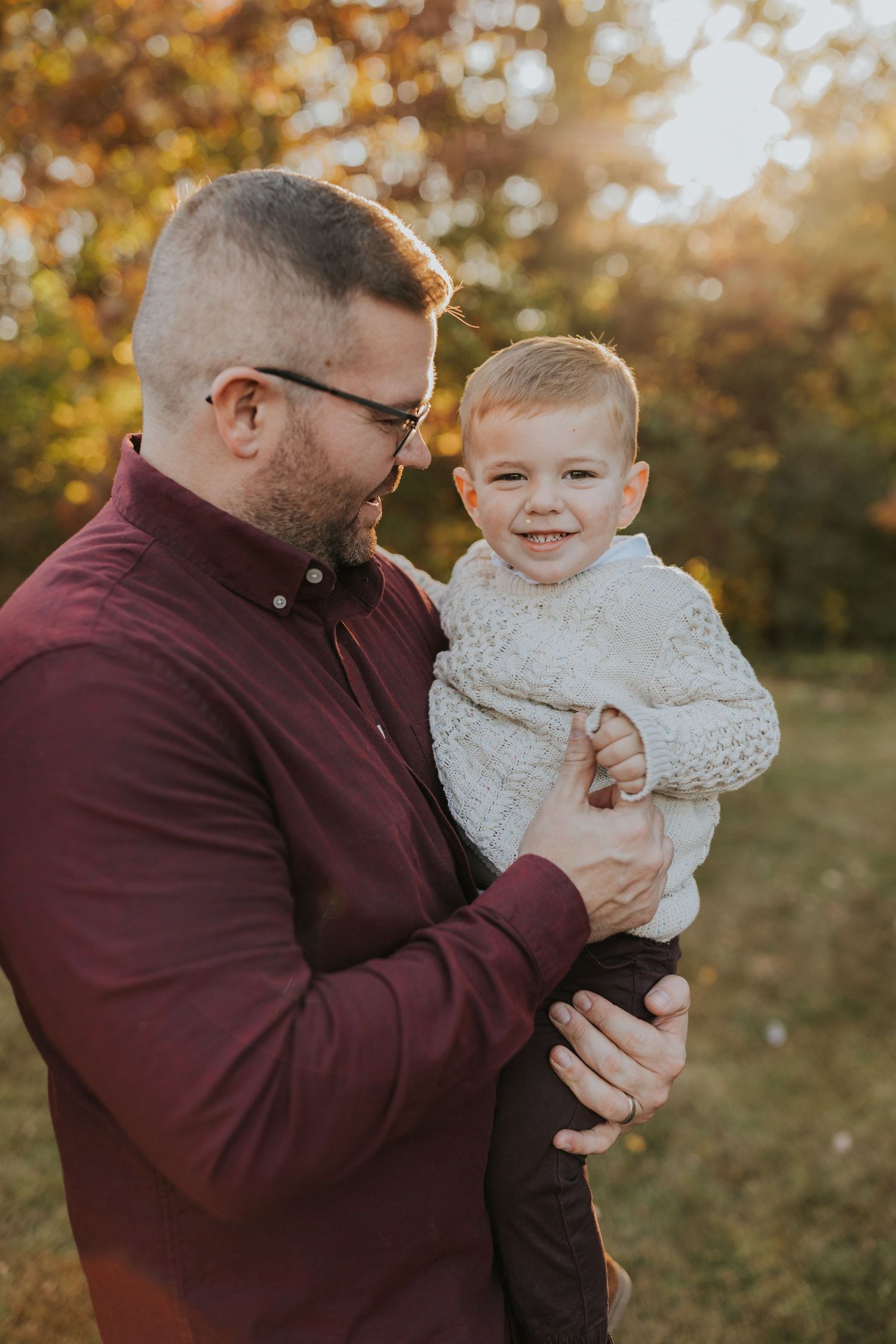 A father holds his smiling toddler son in golden autumn sunlight during a fall family session.