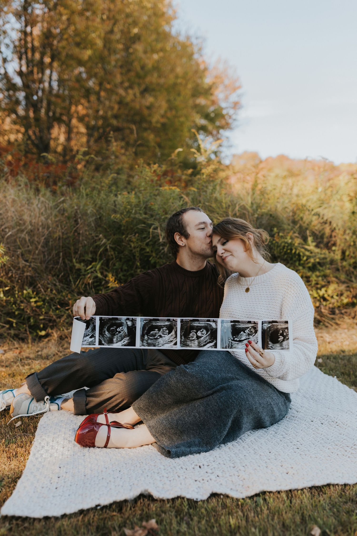 Couple sitting on blanket kissing while displaying ultrasound photos during golden hour fall maternity shoot.