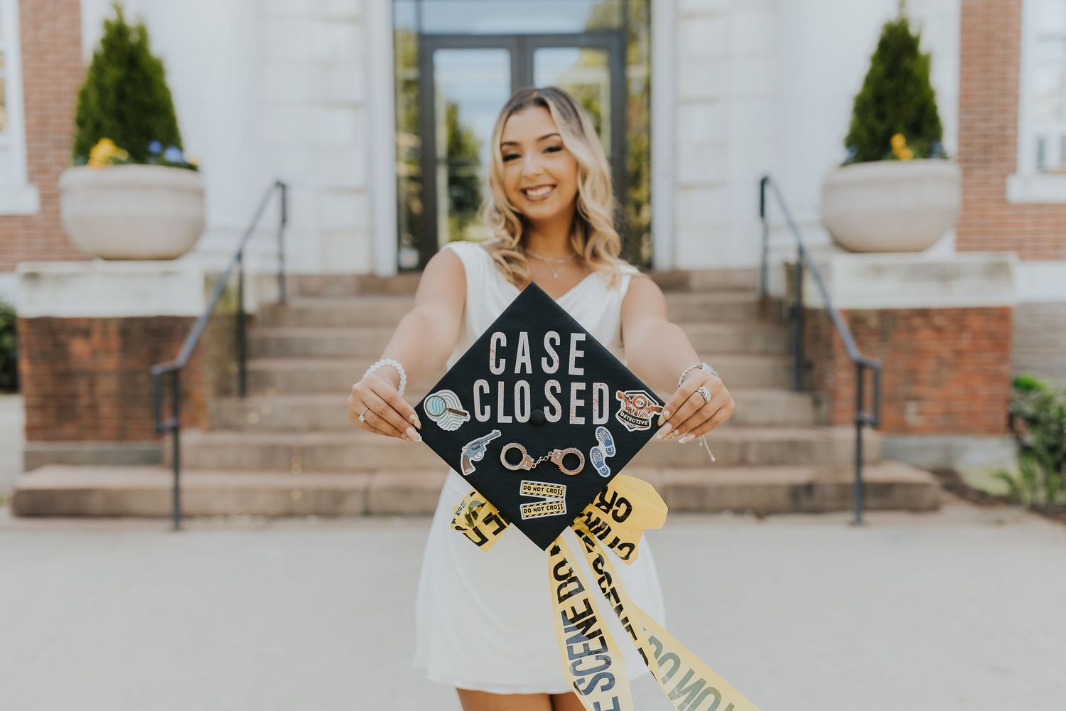 Smiling female graduate holding decorated Case Closed graduation cap with crime scene tape sash.