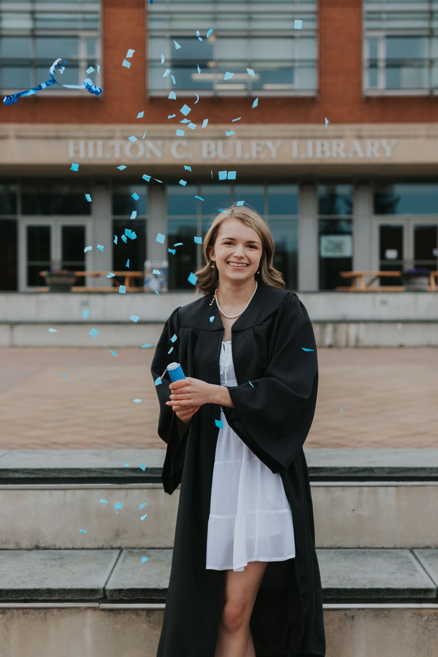 Smiling graduate in black gown celebrates with blue confetti outside Hilton C. Buley Library.