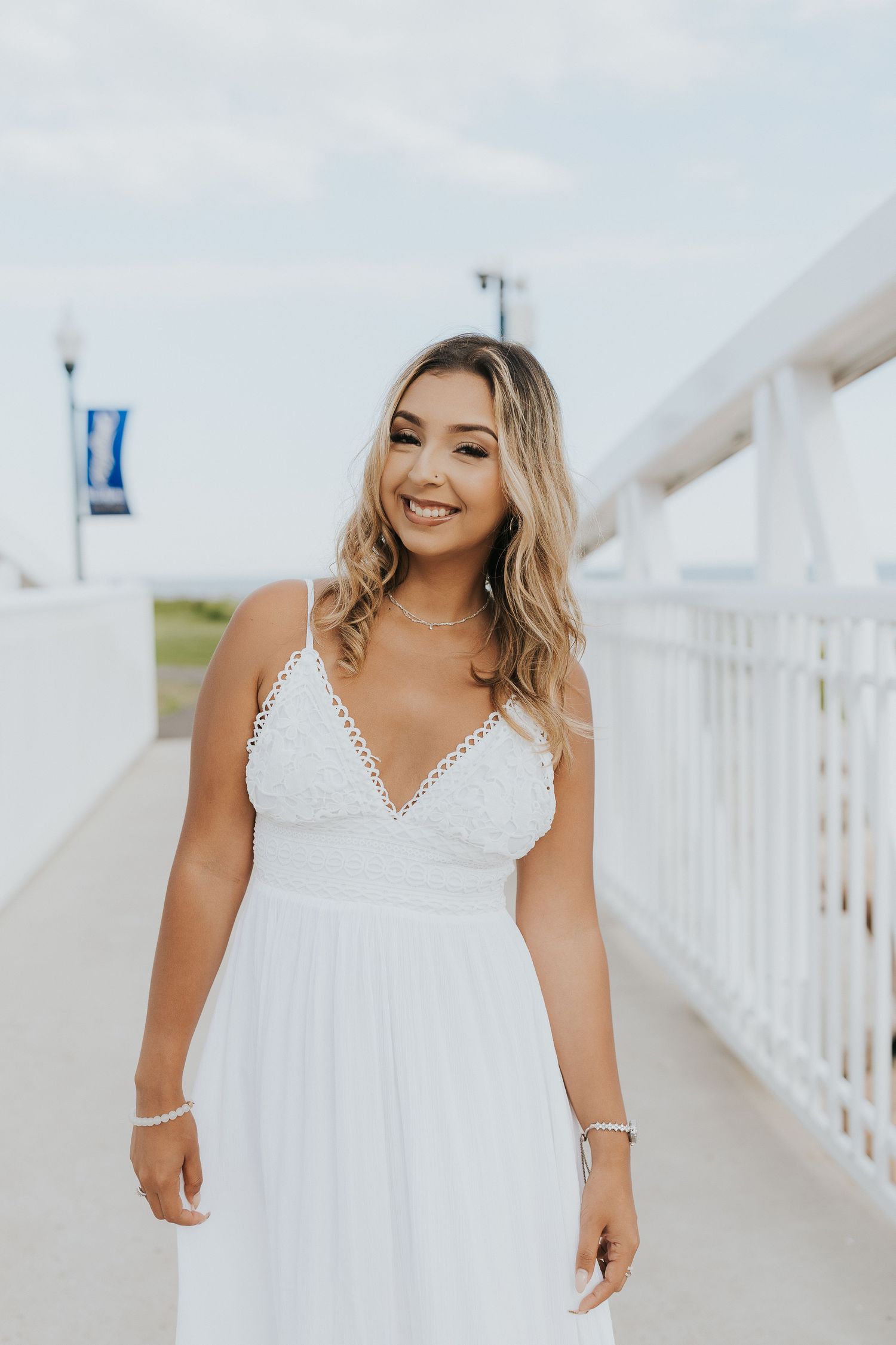 Smiling young woman in white spaghetti strap dress walks along white railing bridge with blue university banners.