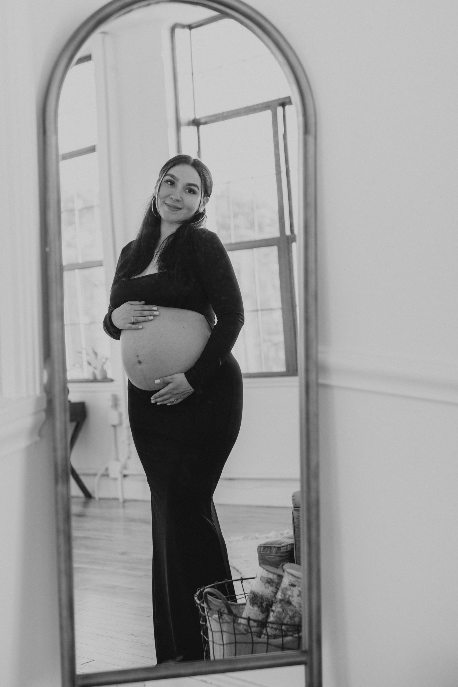Black and white reflection of smiling pregnant woman in black dress holding her belly.