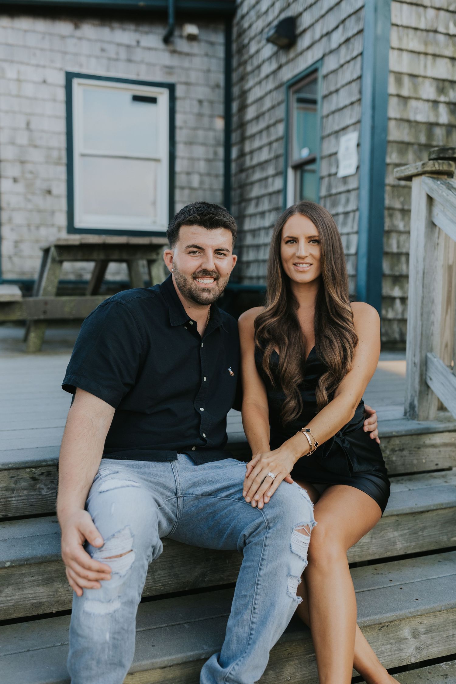 Smiling couple sitting on wooden steps outside rustic building, casual engagement or couples portrait.