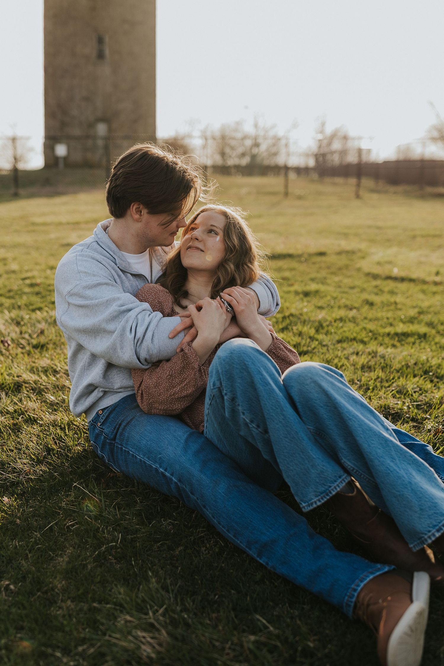 Couple sits together on grass at sunset near a stone tower in a romantic embrace.