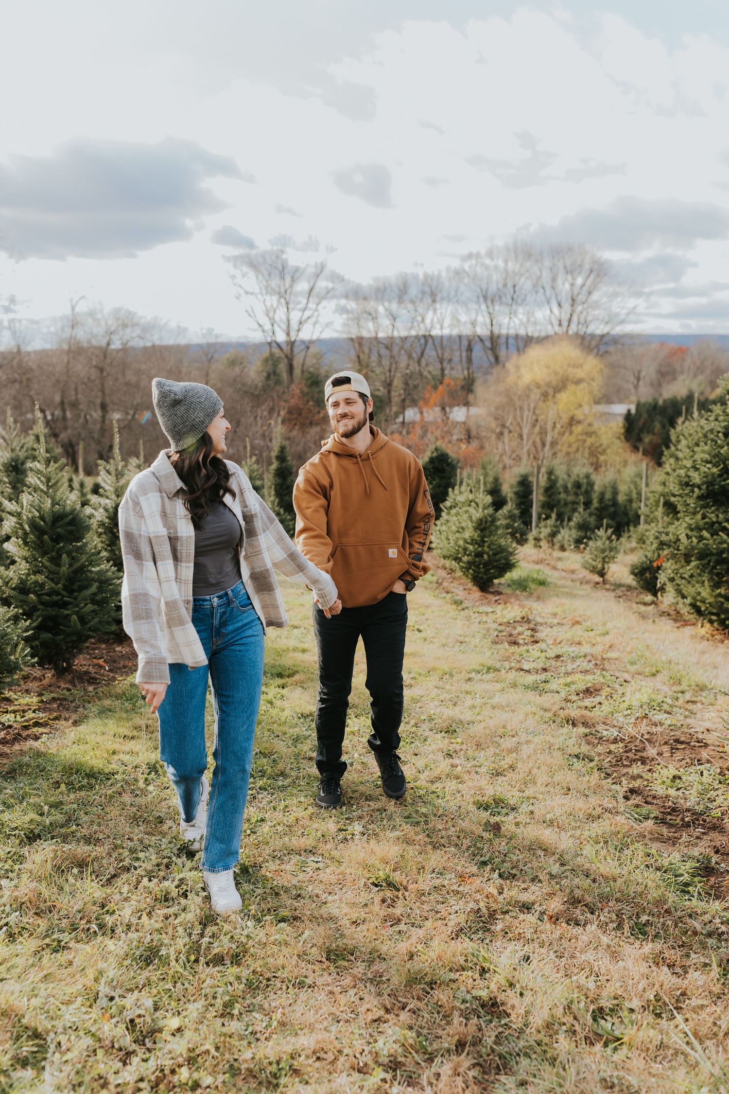 Couple walking hand in hand through a Christmas tree farm on a cloudy day.