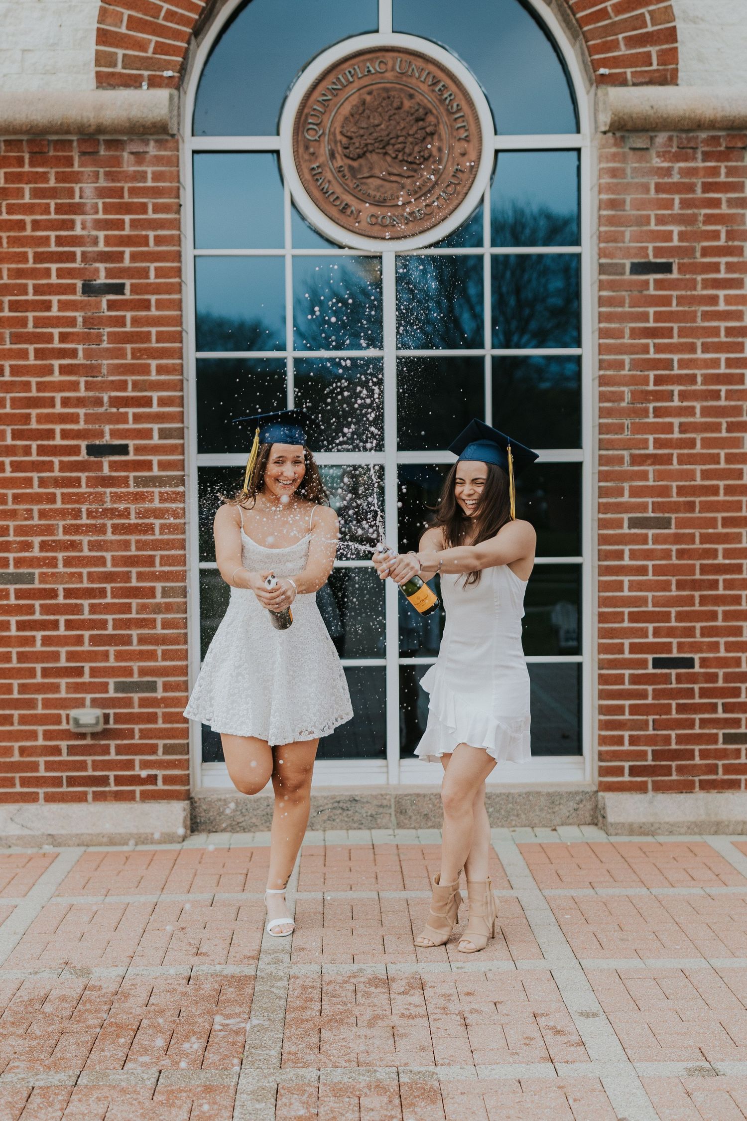 Two graduates in white dresses and caps celebrating with champagne spray outside brick building.