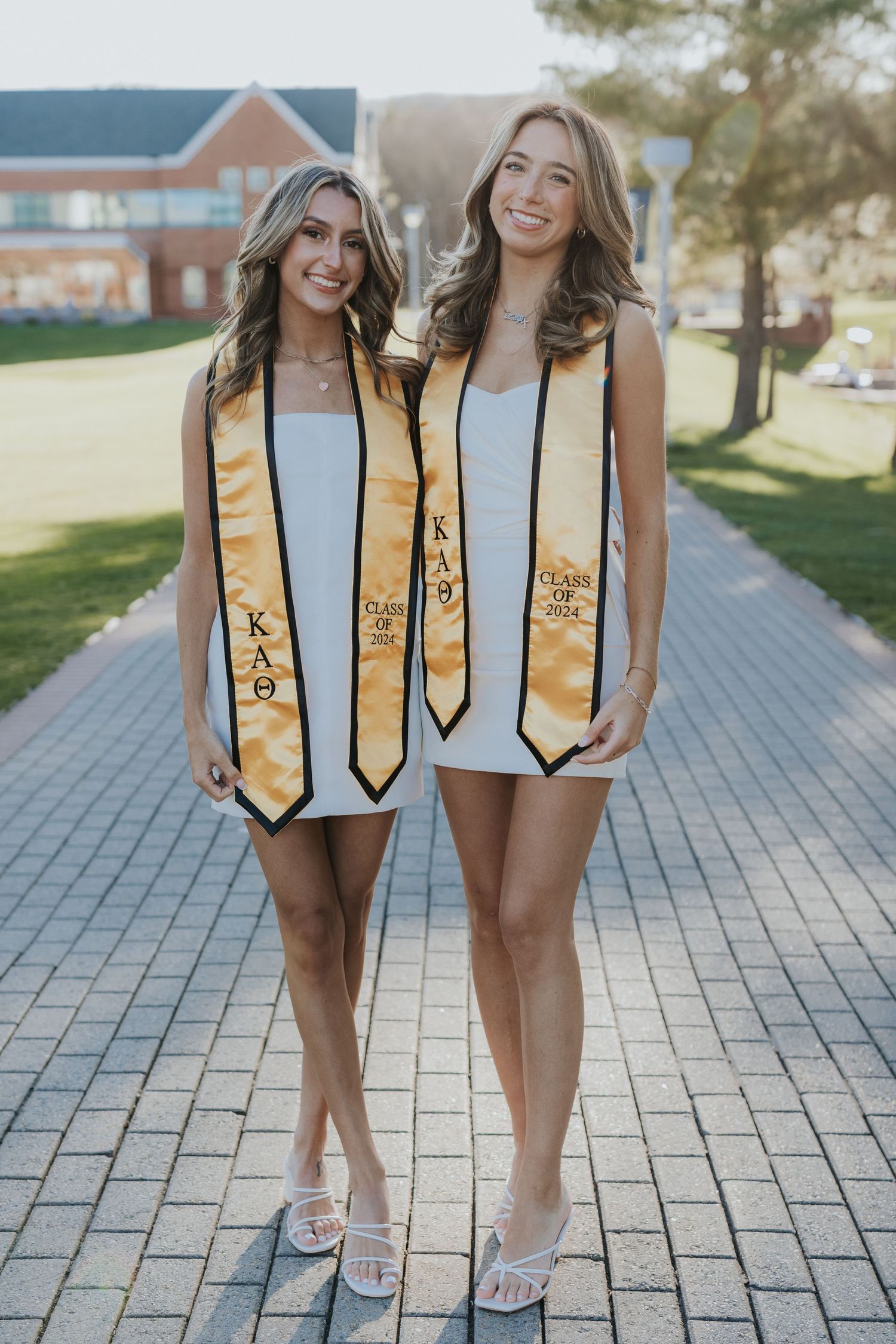 Two smiling women in white dresses wearing Kappa Alpha Theta graduation stoles on brick pathway.