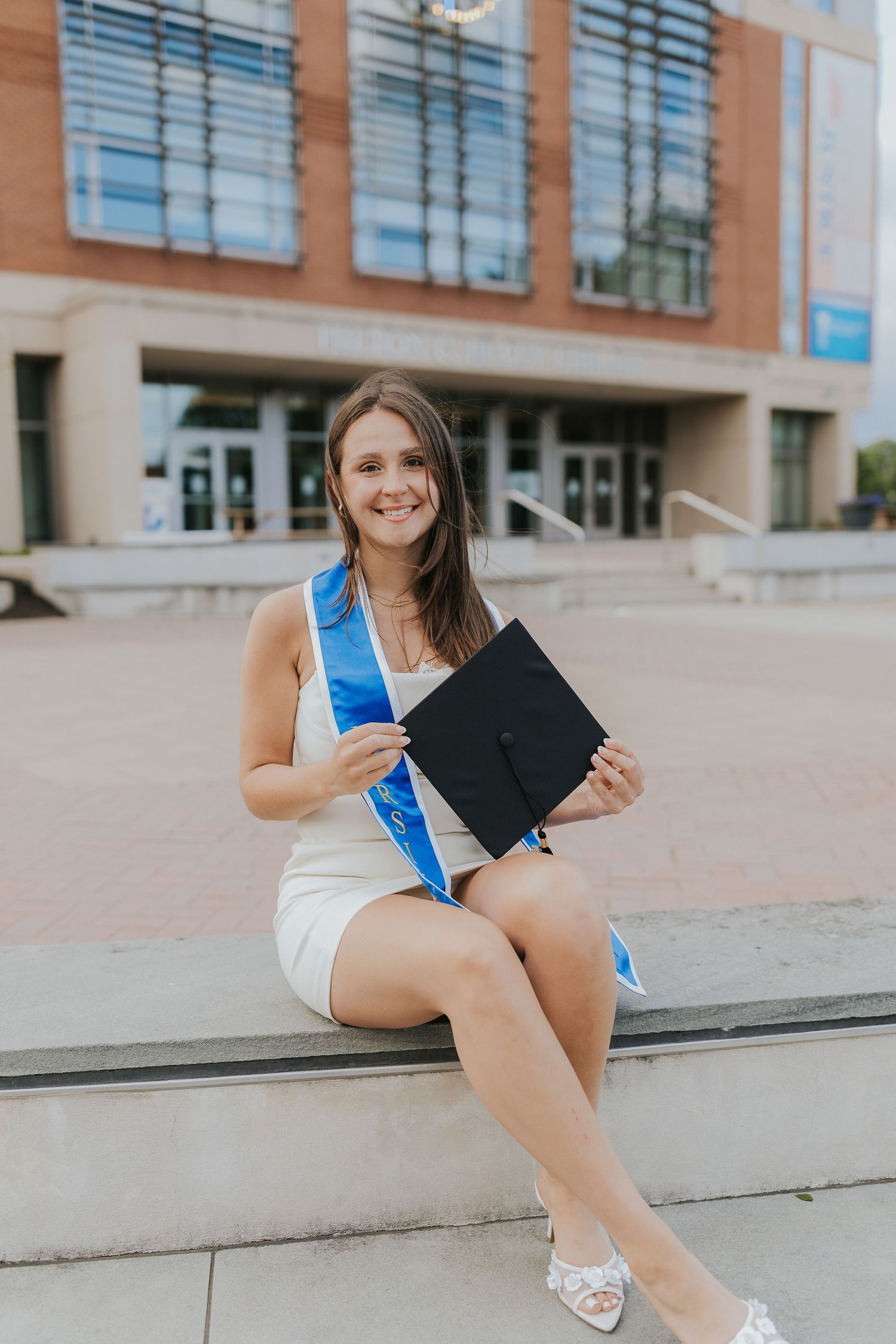 Smiling nursing graduate holds cap while sitting outside campus building.