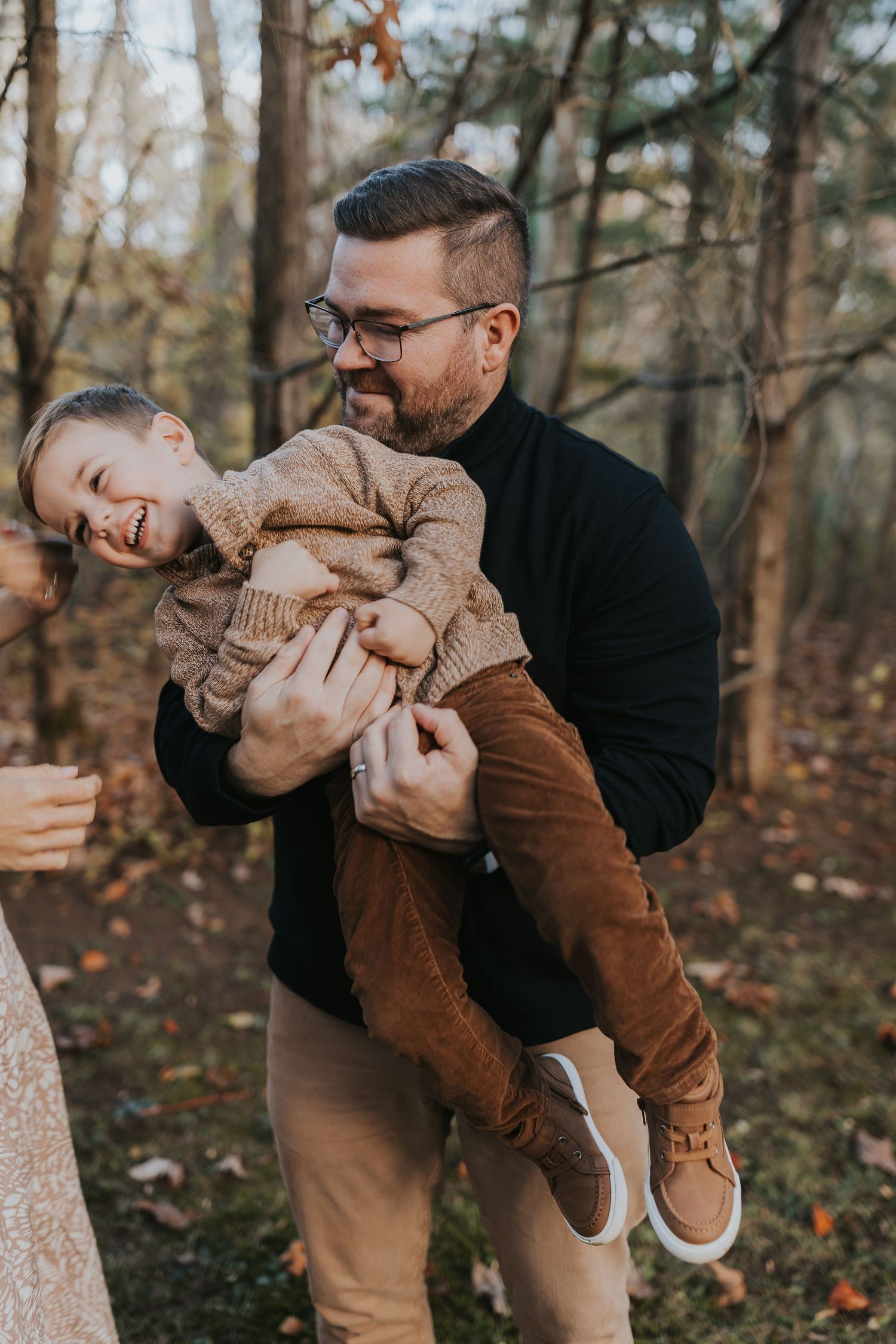 Father laughing while playfully holding his giggling young son in an autumn forest setting.