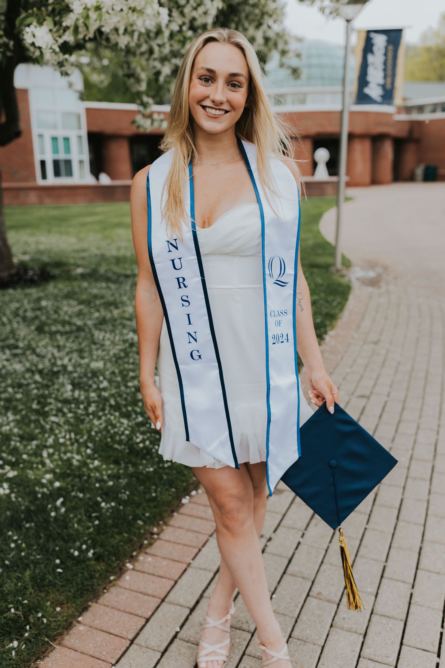 Nursing Class of 2024 graduate in white dress holding navy cap on a blooming campus path.