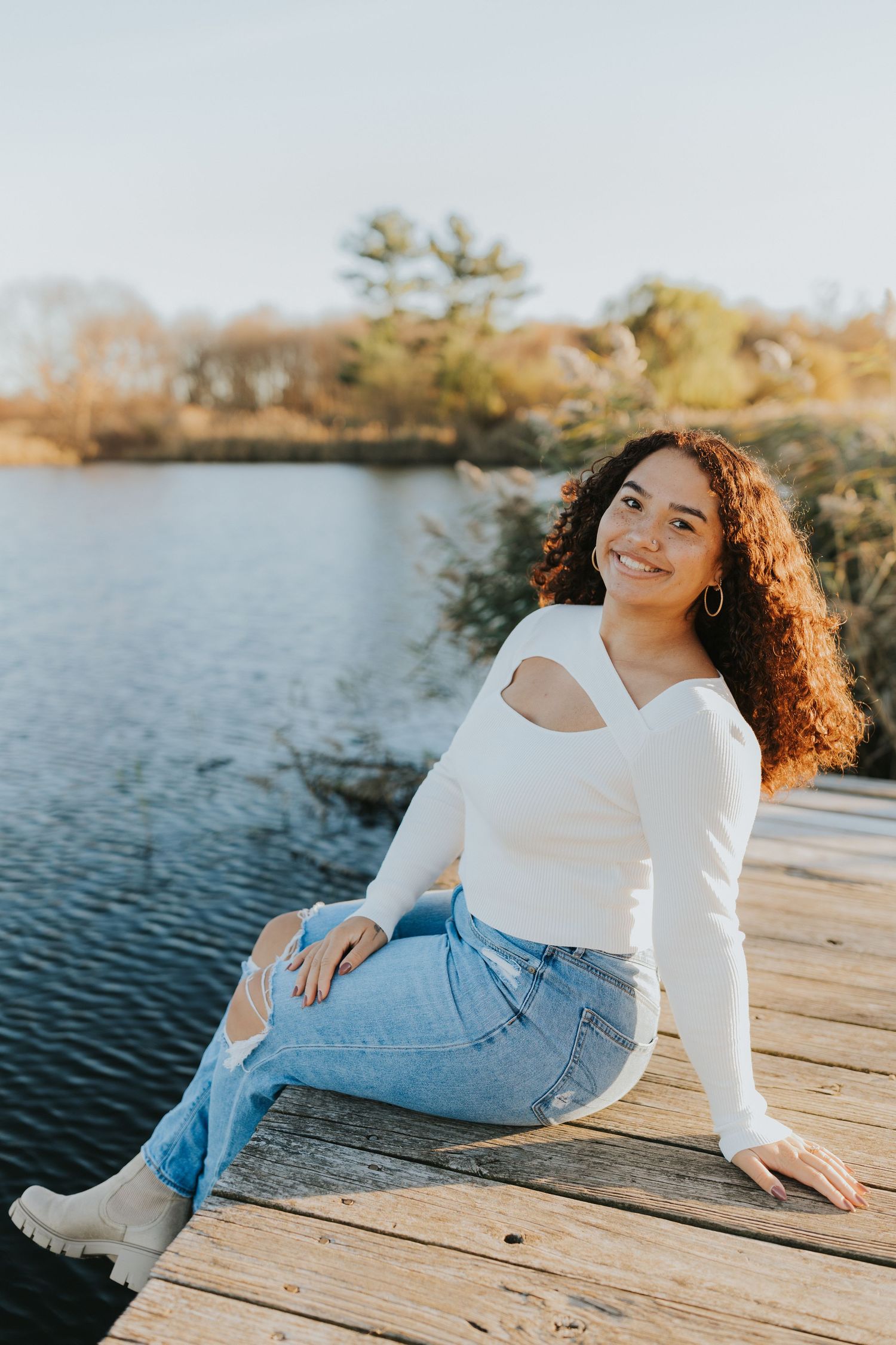 Smiling young woman in white cutout top and ripped jeans sitting on wooden dock by lake.