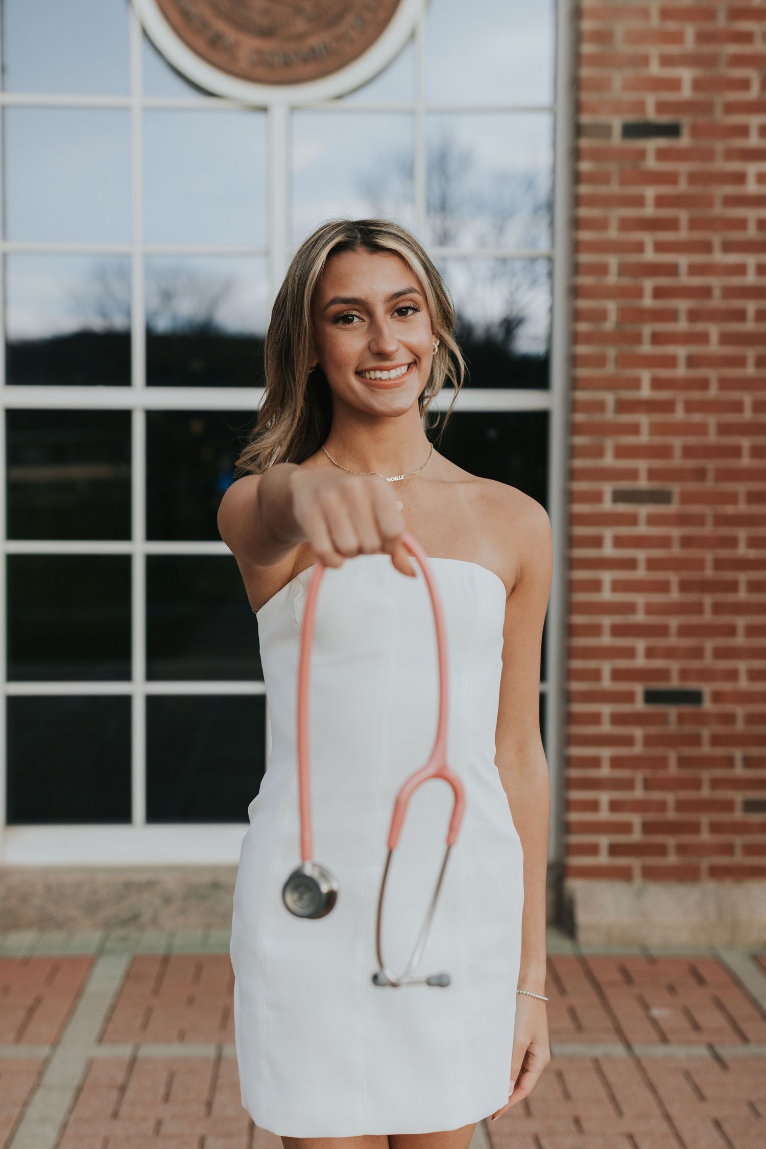 Nursing graduate in white dress holds pink stethoscope toward camera outside brick university building.
