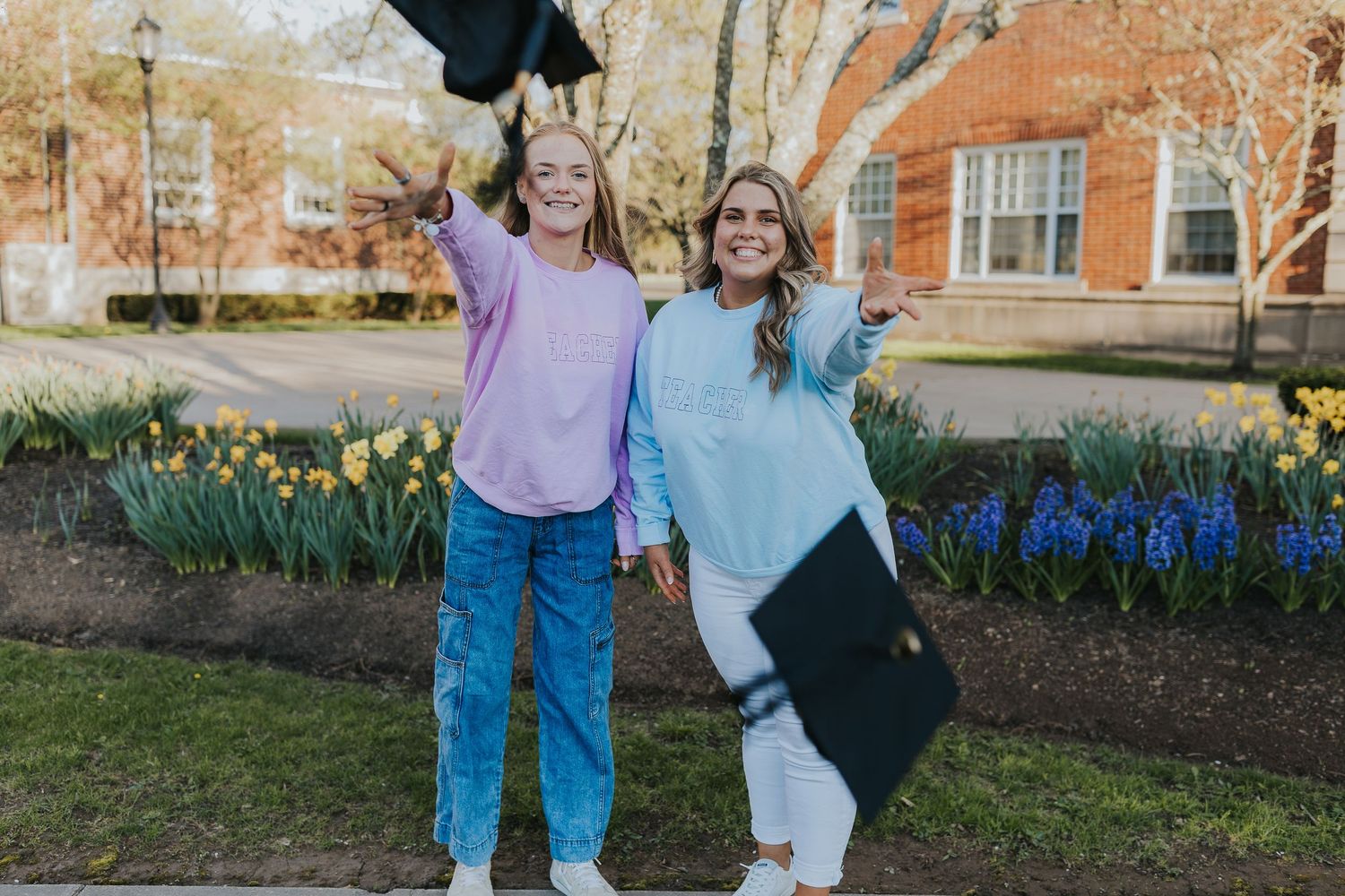 Two smiling women holding graduation caps near colorful spring flowers on university campus.