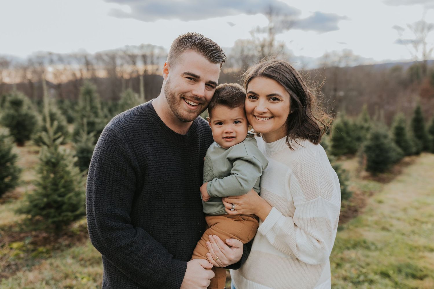 A smiling young couple holds their baby boy at a Christmas tree farm during a fall family photo session.