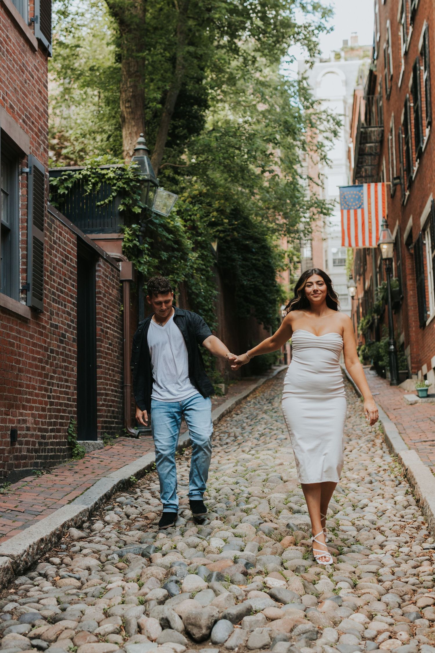 Couple holding hands and walking down a historic cobblestone street lined with brick buildings and greenery.