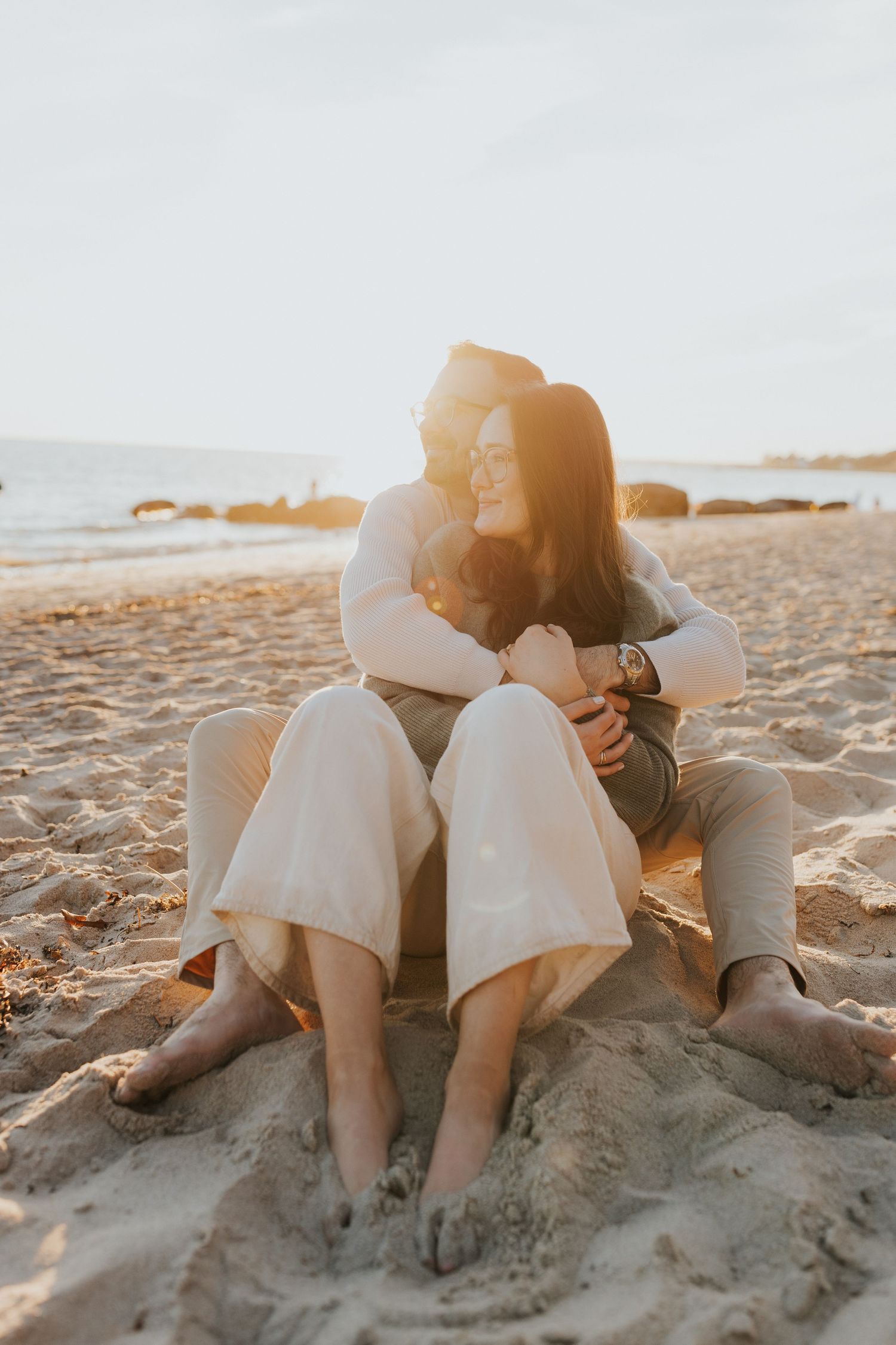 Couple sitting together on sandy beach at golden hour, woman embracing man from behind in warm sunset light.