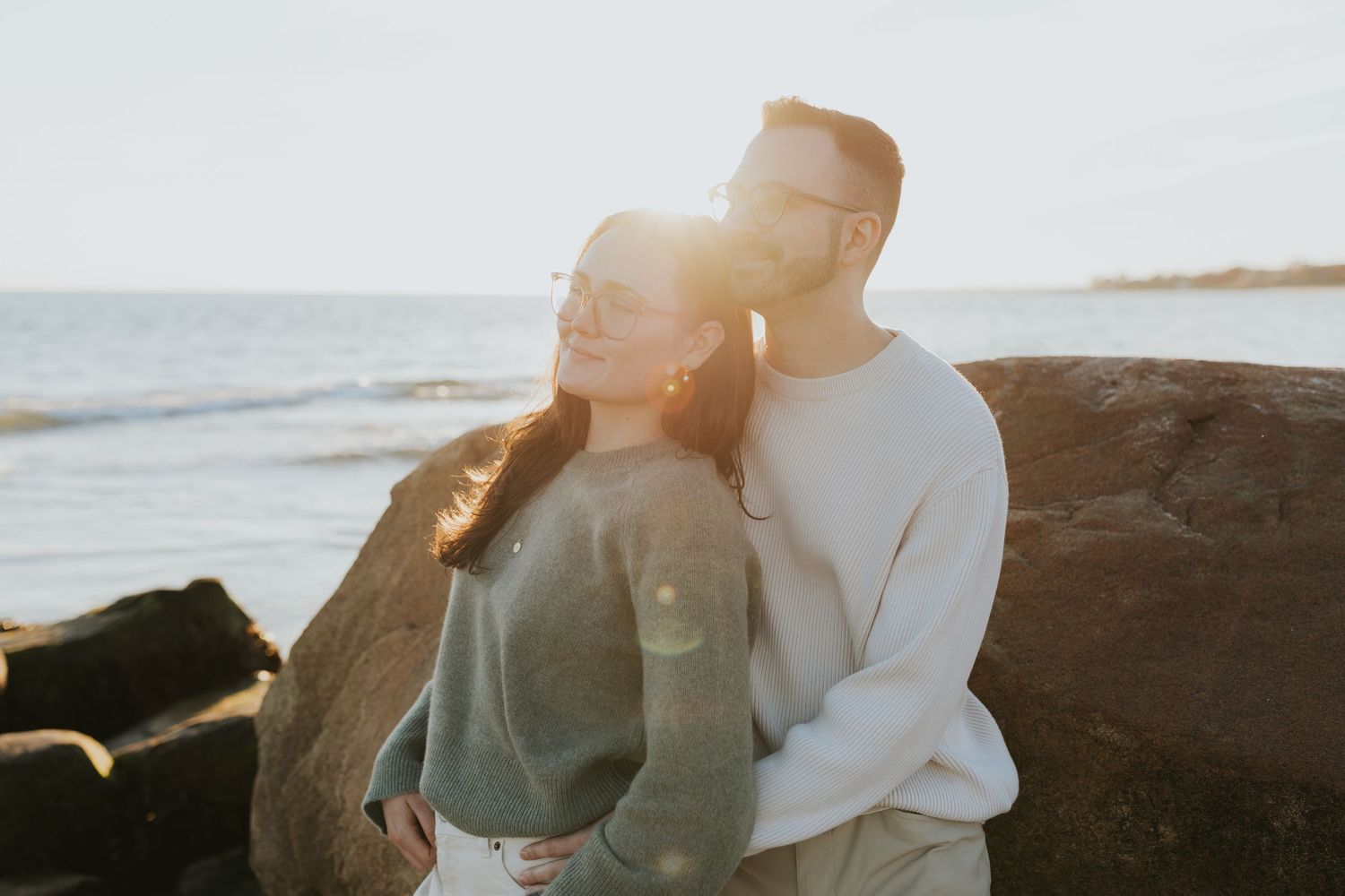 Couple cuddling on rocky beach with warm golden sunset light streaming behind them.