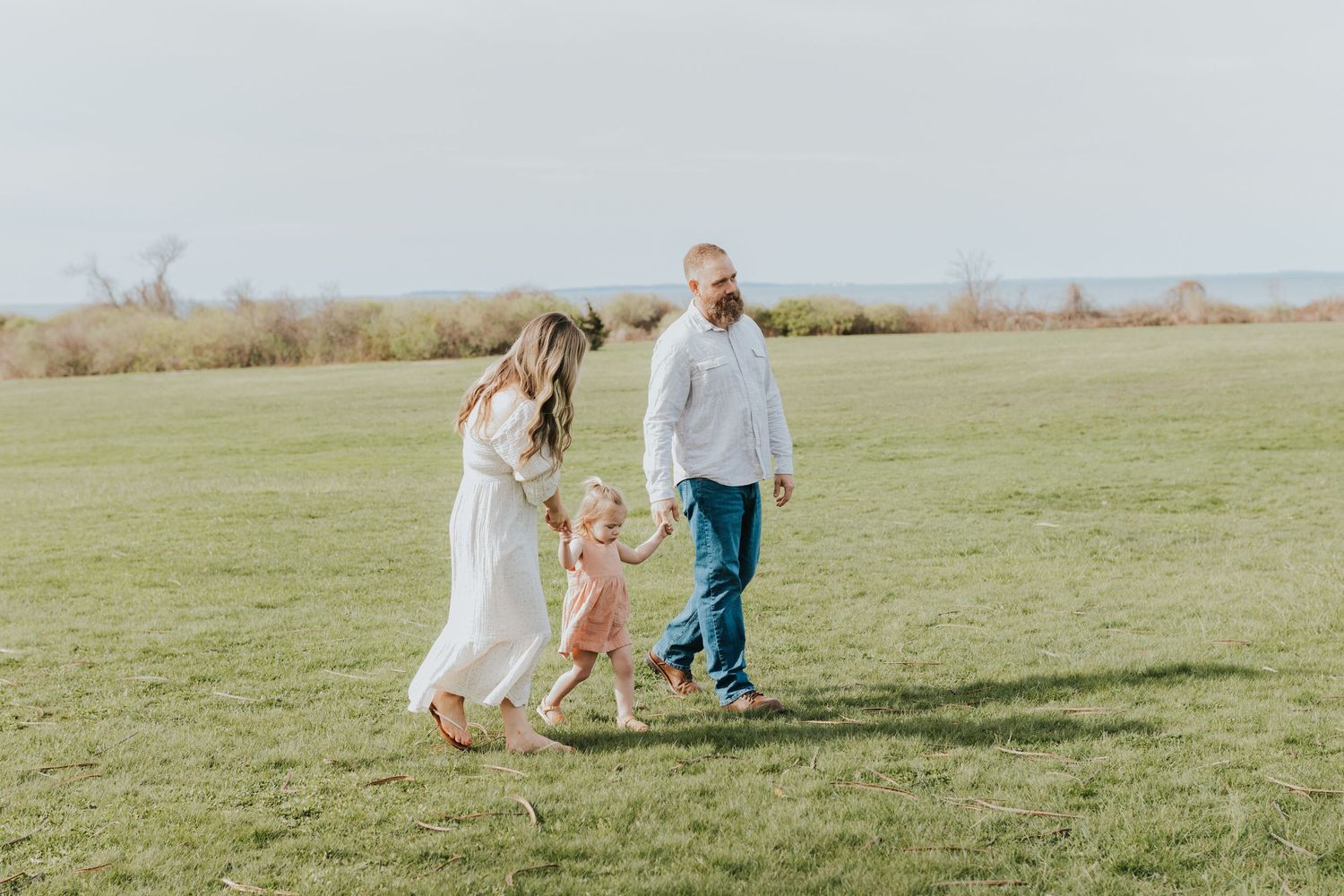 Family of three playing in green field, parents holding toddler girl's hands in white and pink outfits.