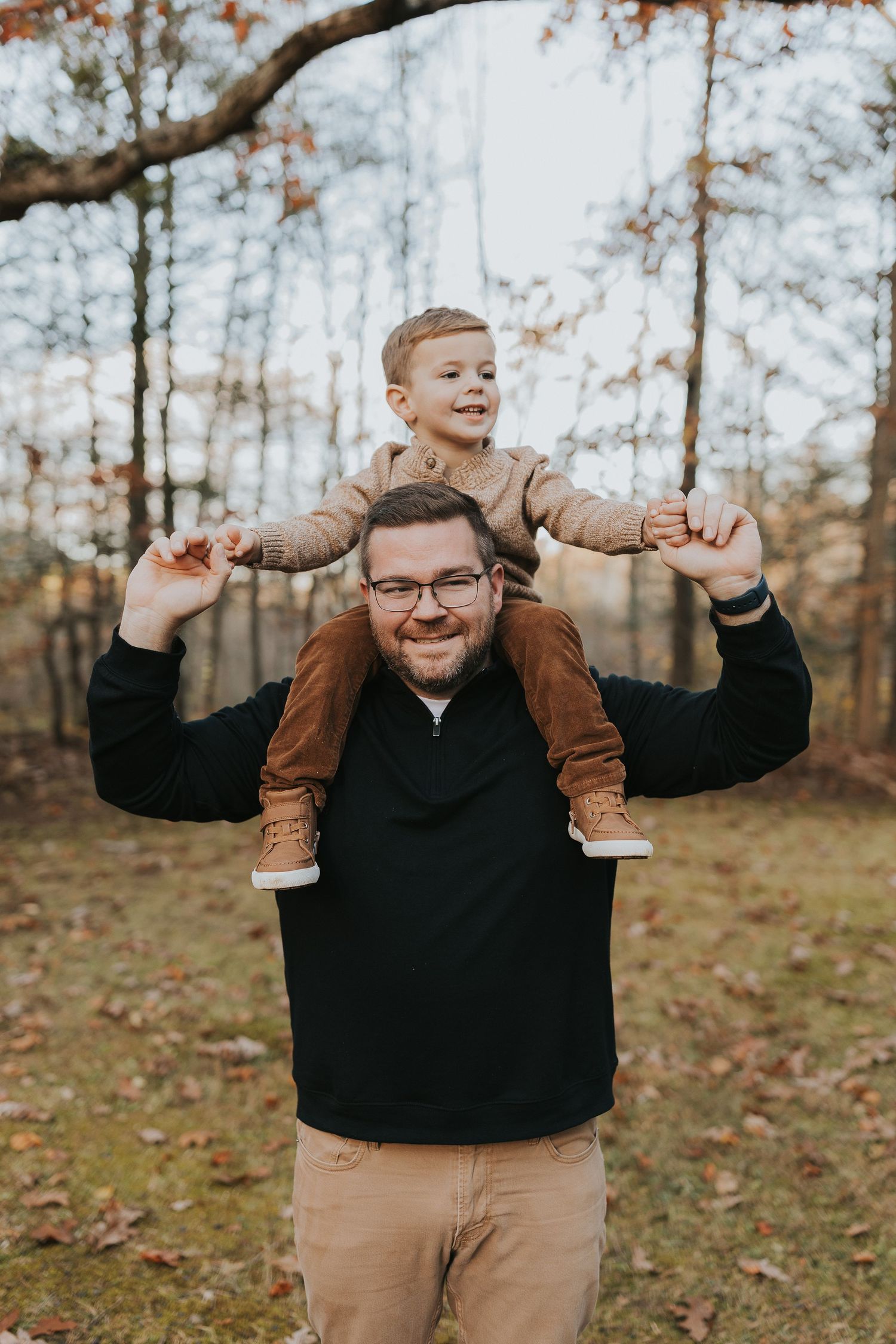 A smiling father carries his happy toddler son on his shoulders in an autumn forest setting.