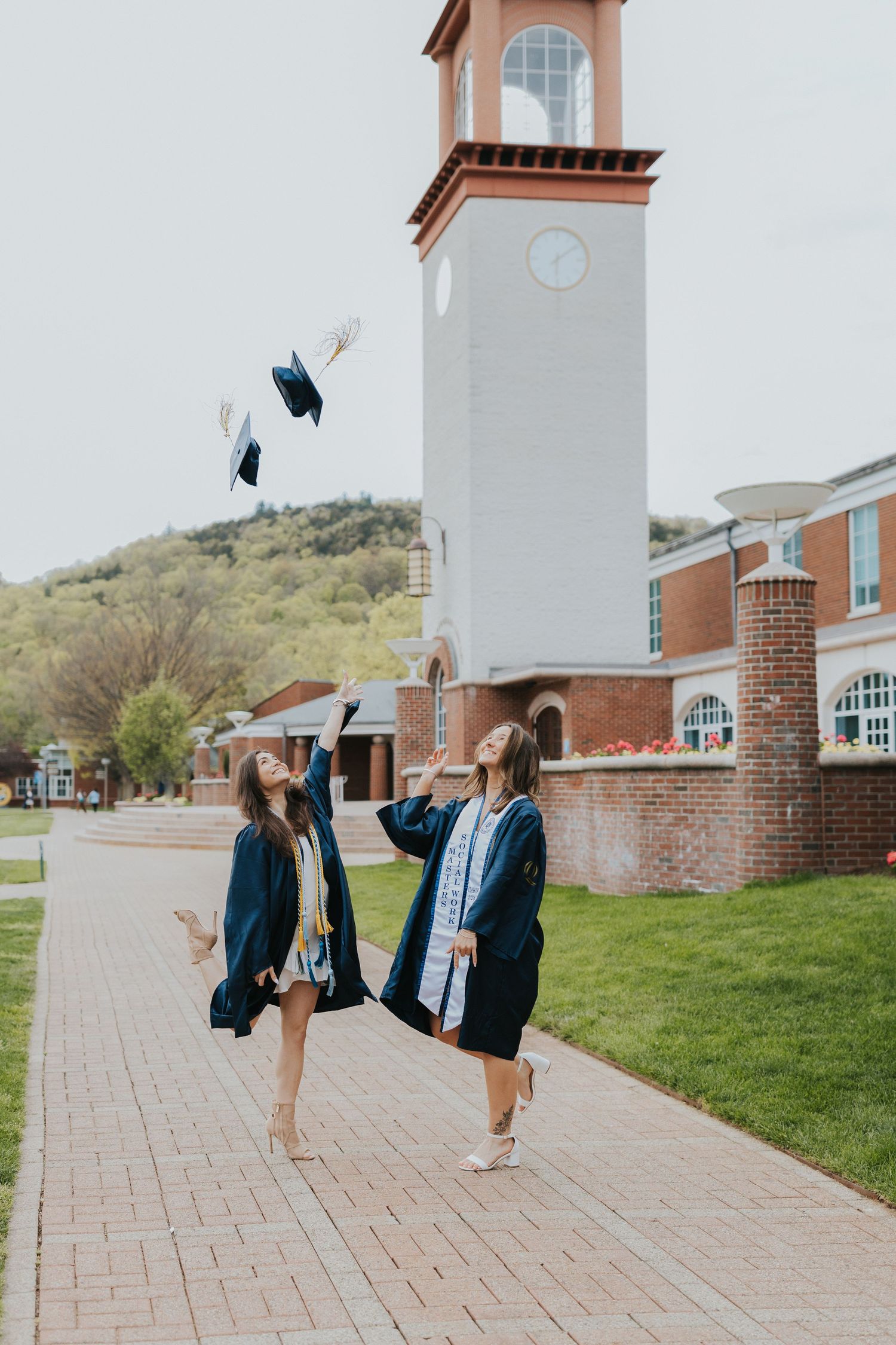 Two graduates in navy gowns toss caps in air near brick clock tower building on college campus.