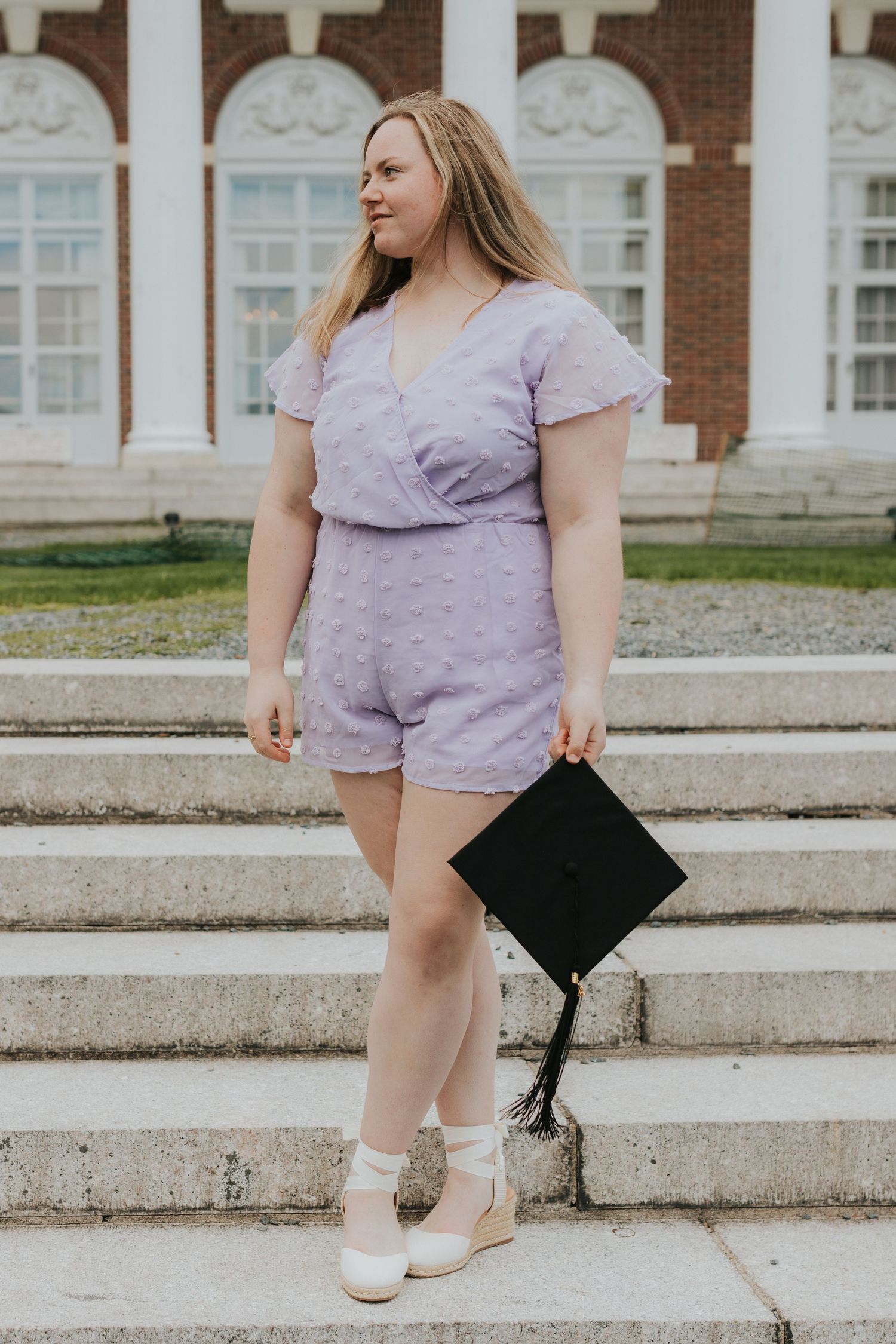 Young woman in lavender romper holds graduation cap while posing on campus steps.