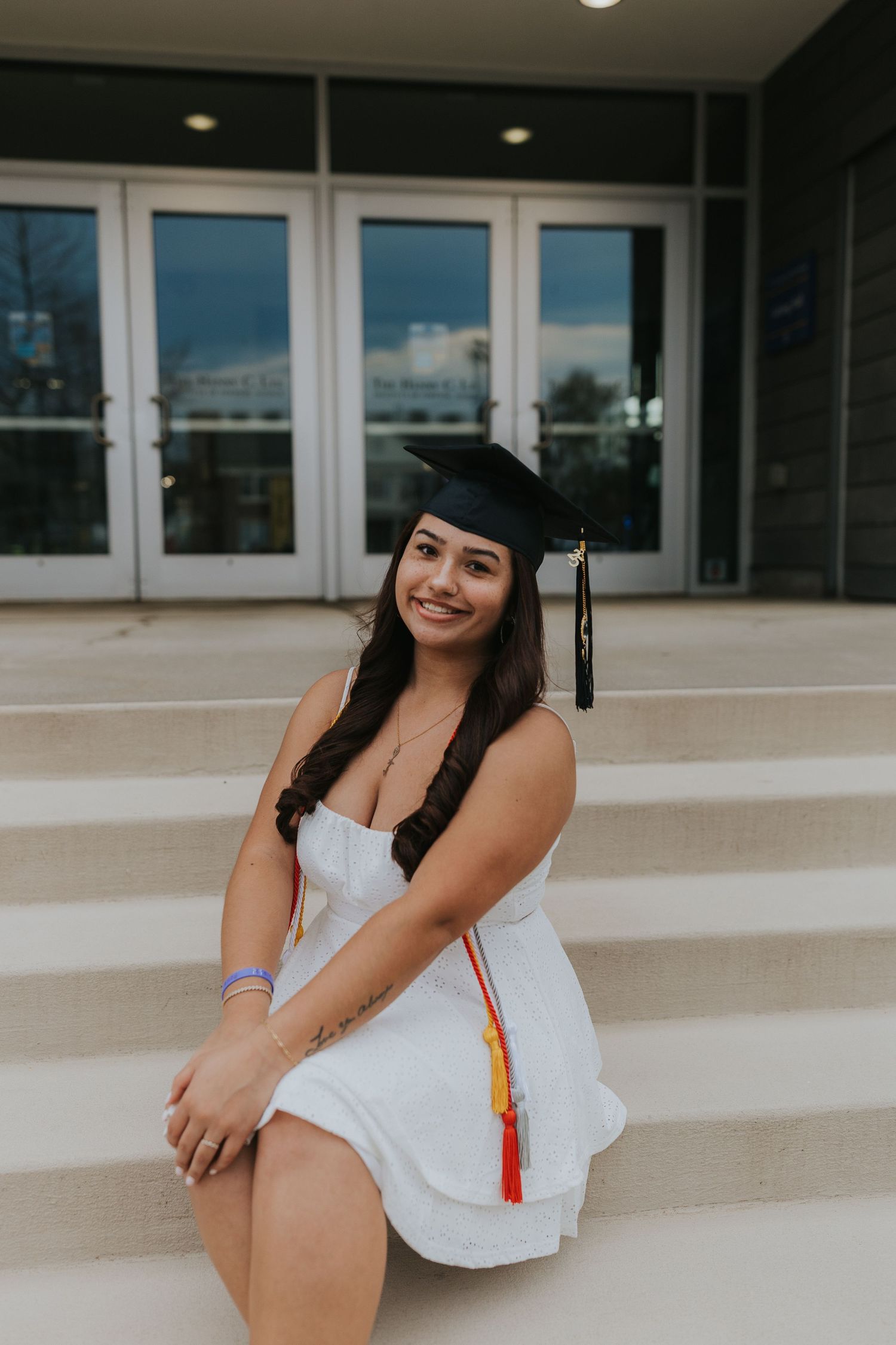 Smiling graduate in white dress and black cap sits on steps outside campus building.