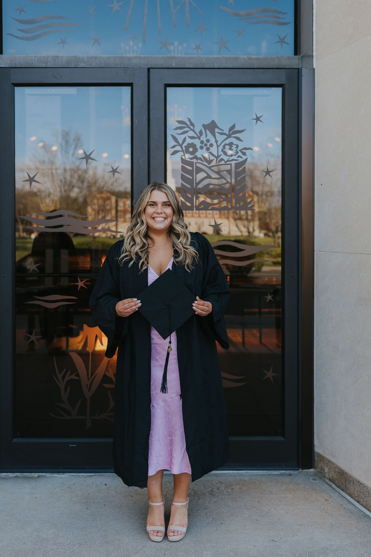 Smiling graduate in black gown and pink dress holding cap outdoors.