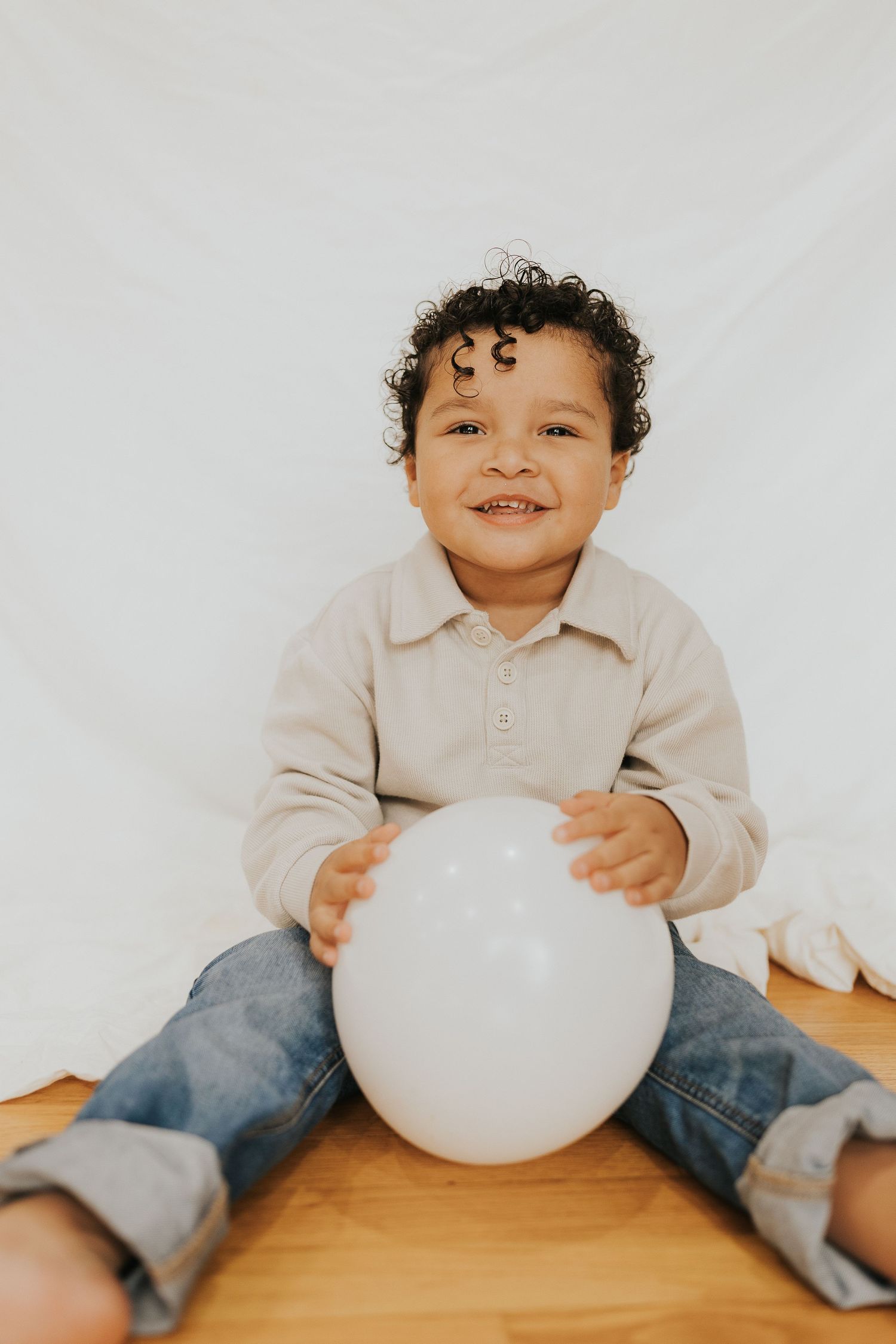 A smiling curly-haired toddler in jeans sits on hardwood floor holding a white balloon indoors.