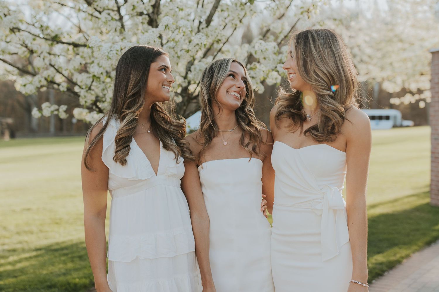 Three smiling women in white dresses standing together near blooming cherry blossom trees outdoors.