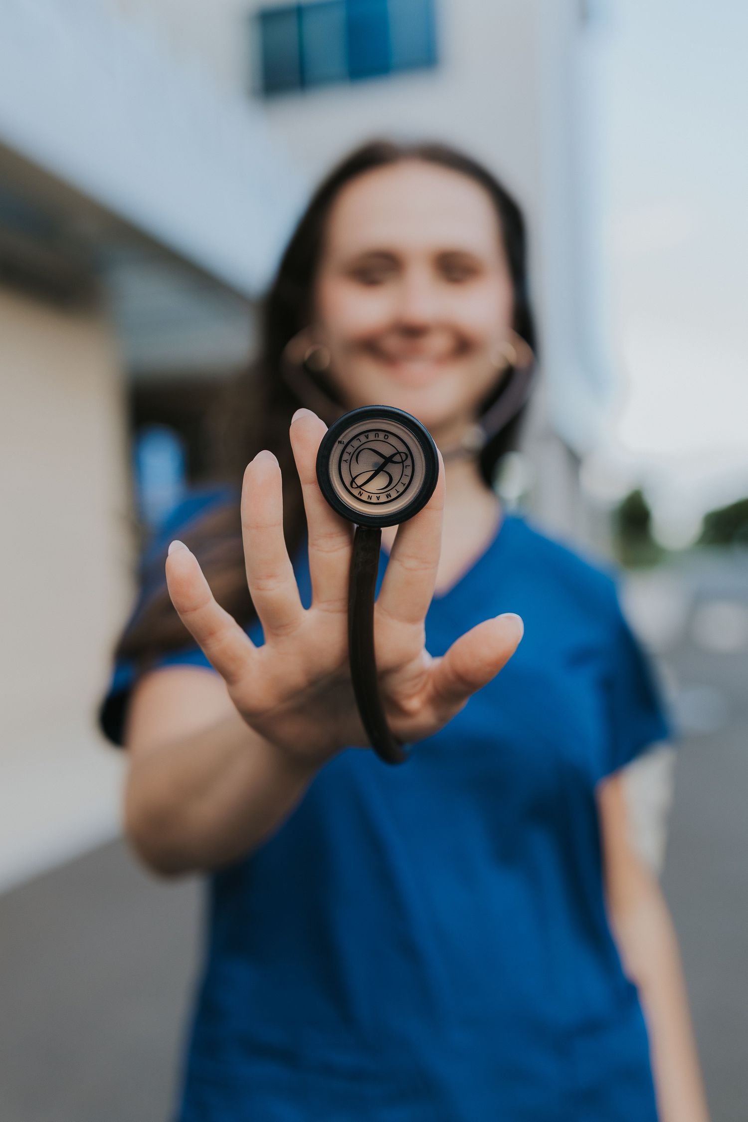Nursing student in blue scrubs holds stethoscope toward camera outdoors.