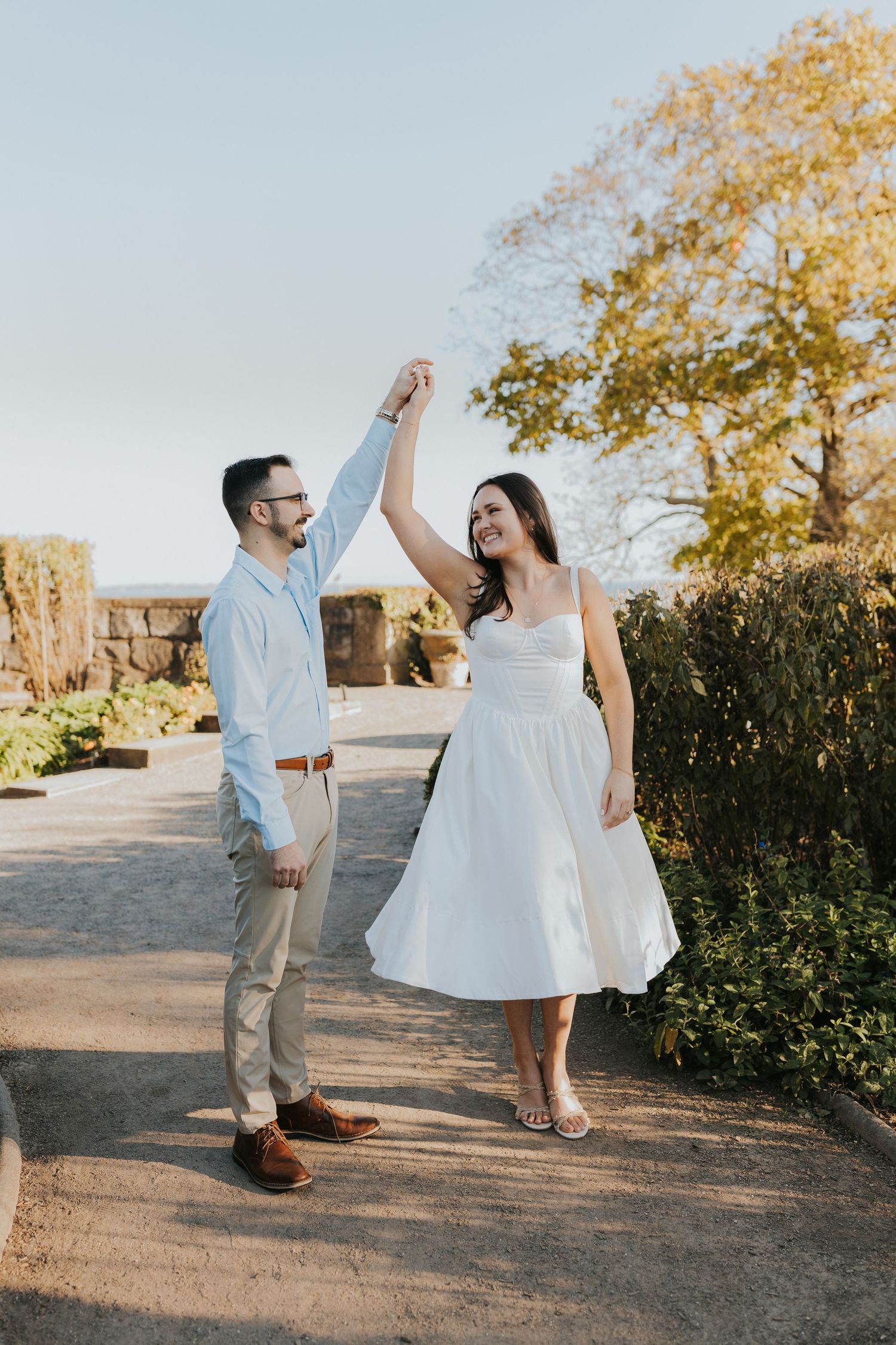 Couple dancing outdoors in fall, woman in white dress twirling, man in light blue shirt smiling.