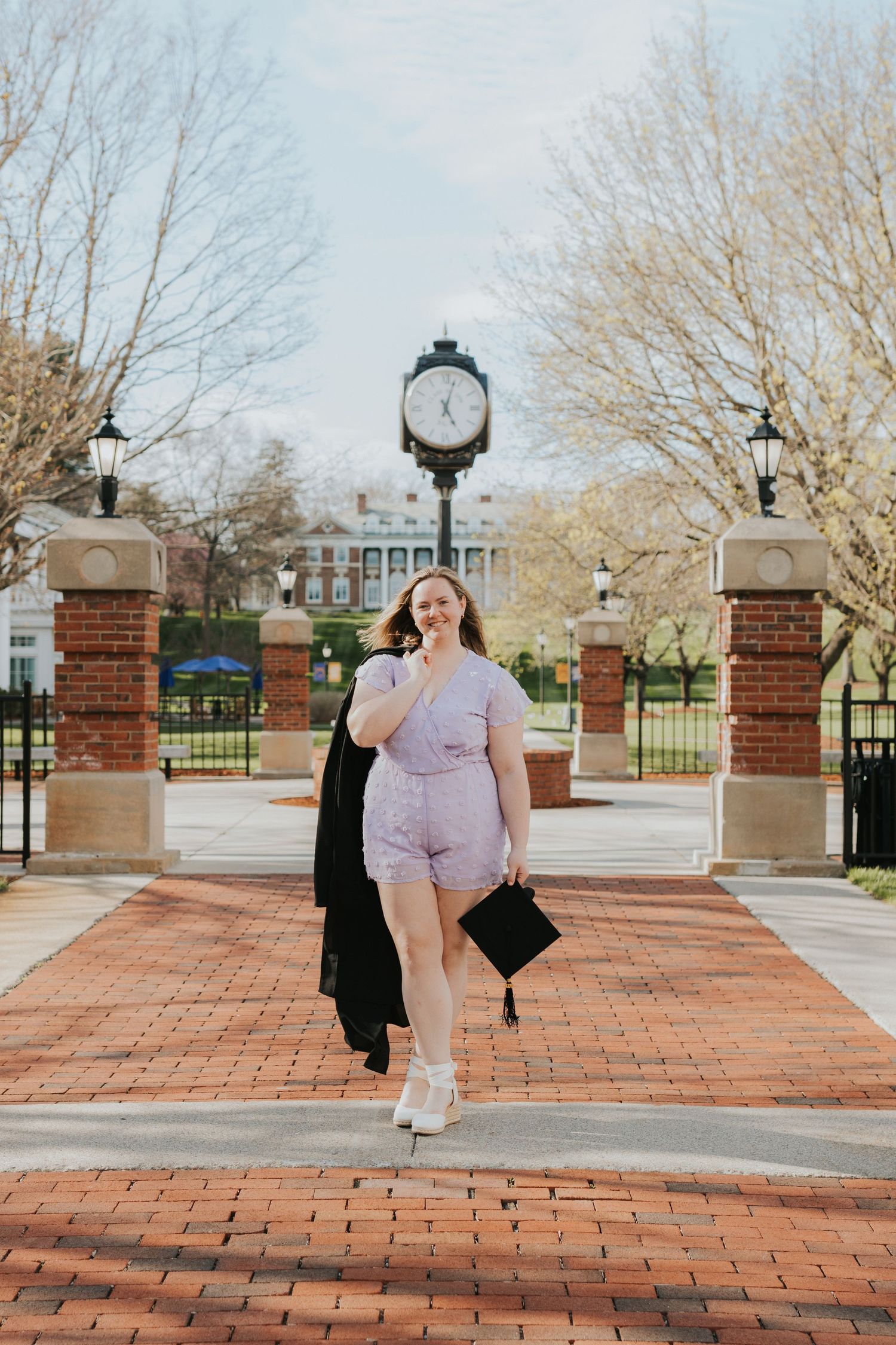 Young woman in lavender romper holding graduation cap walks brick path with campus clock tower backdrop.
