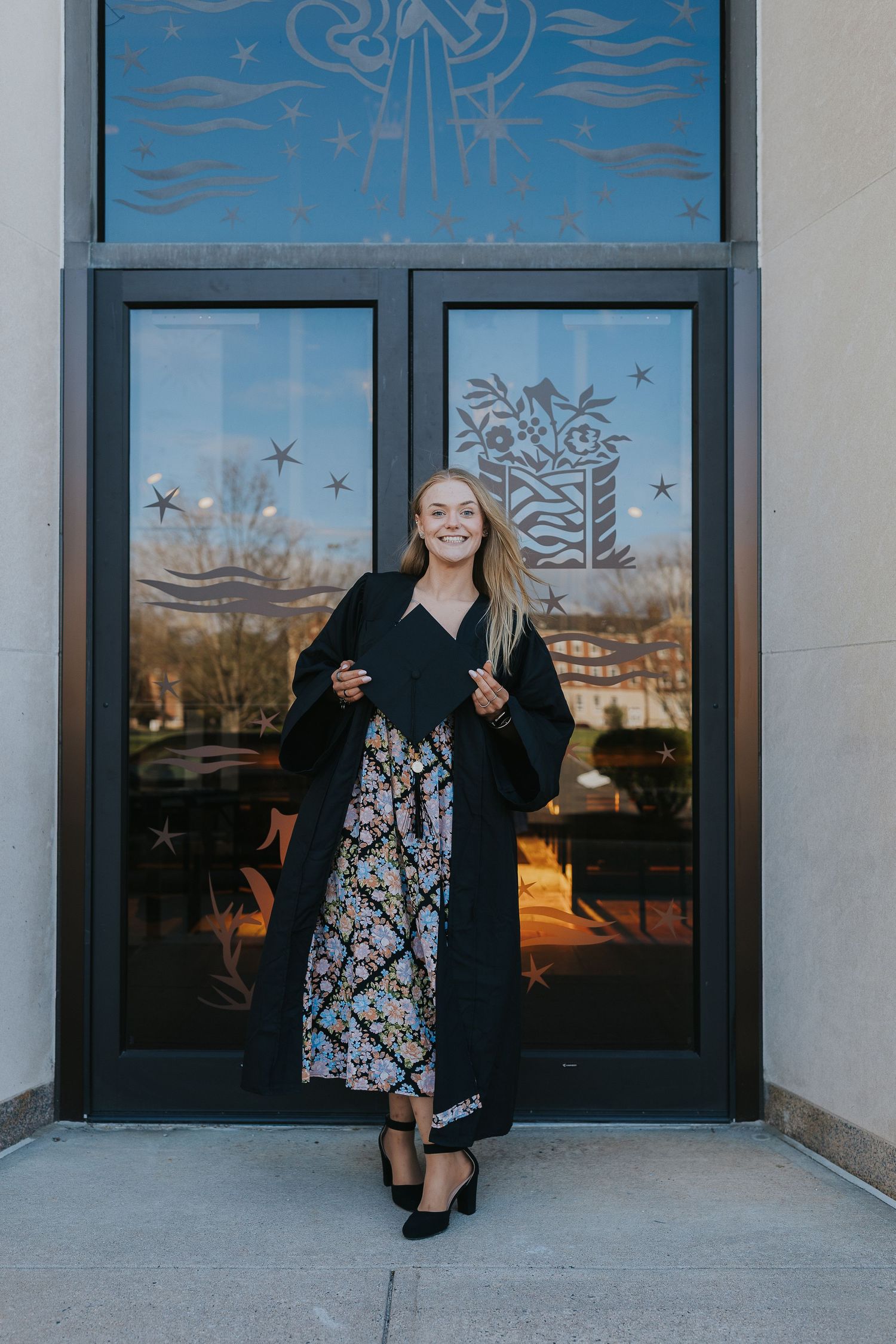 Smiling graduate in black gown over floral dress outside building.