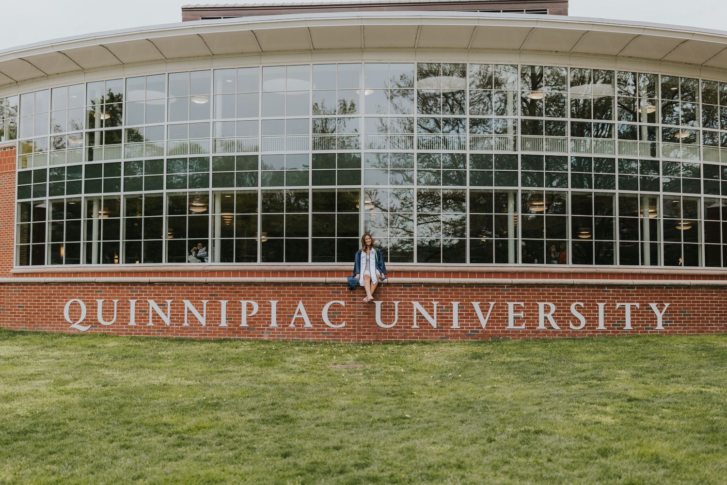 Graduate in cap and gown poses in front of large Quinnipiac University glass building on campus.