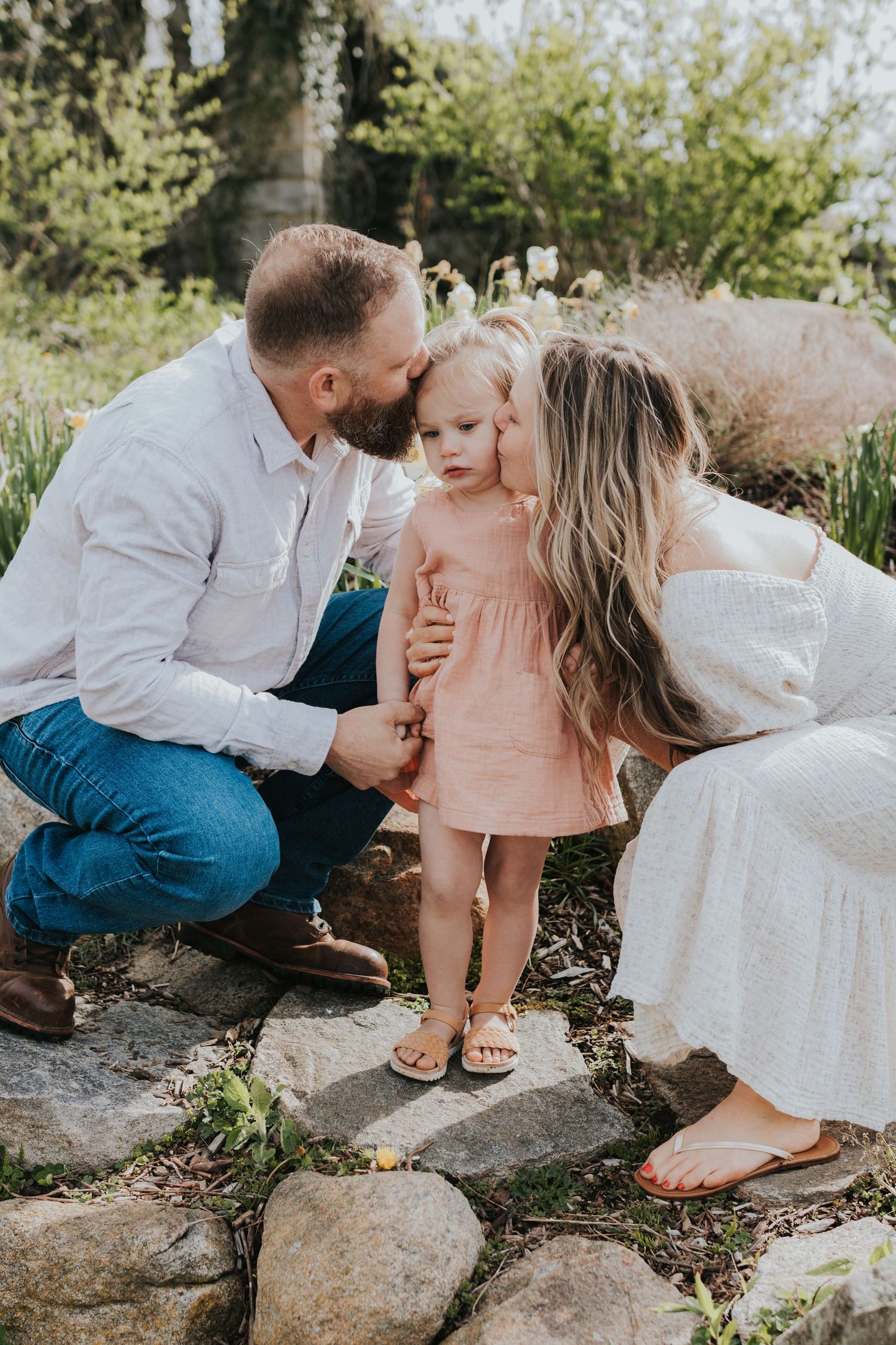 Parents kissing toddler girl in pink dress on rocky garden path surrounded by spring flowers.
