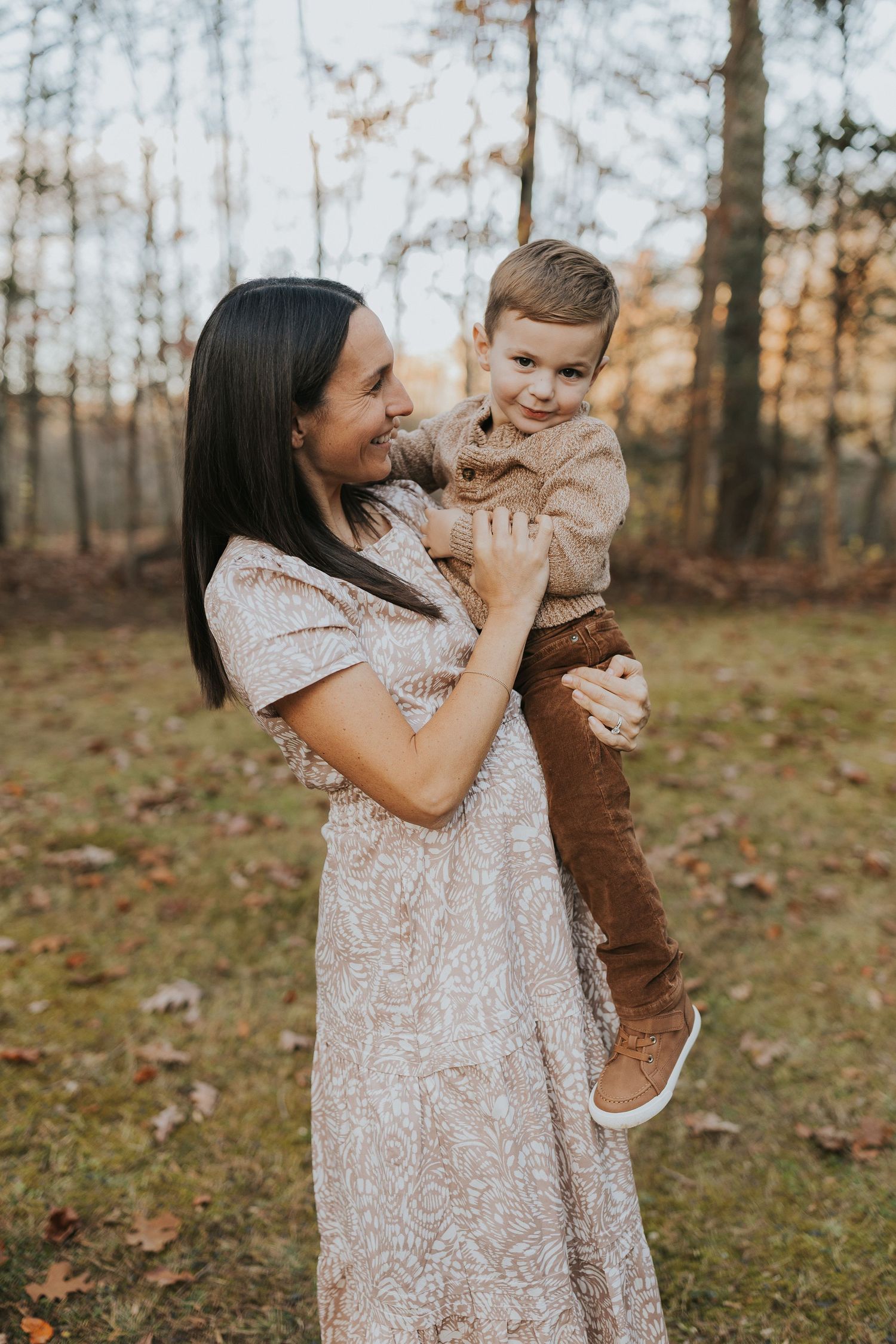 A smiling mother holds her laughing toddler son in a warm autumn forest at golden hour.