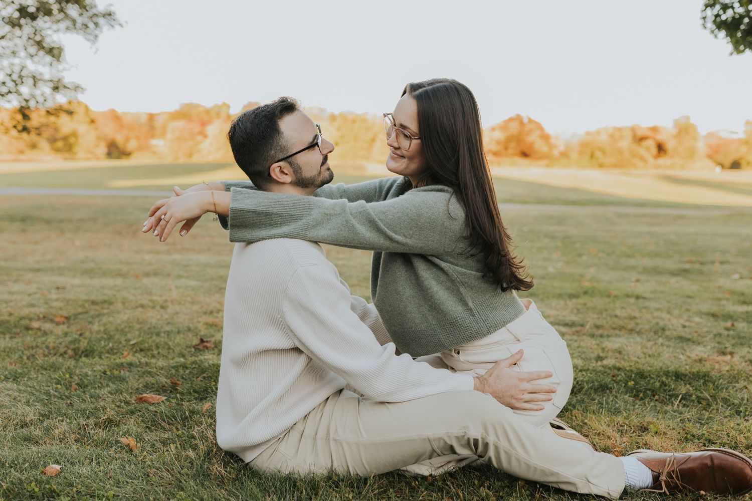Happy couple sitting on grass in autumn park, embracing and gazing at each other lovingly.