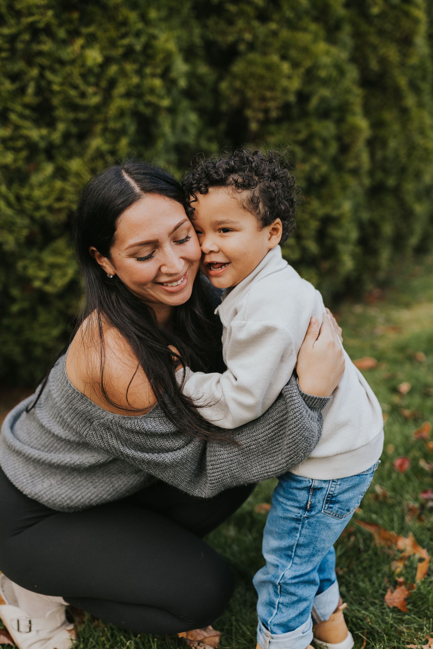 Mother hugging her curly-haired toddler son outdoors in a lush green garden setting.