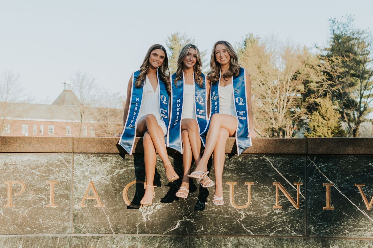 Three smiling female graduates in white dresses and blue stoles sit on a university monument wall outdoors.