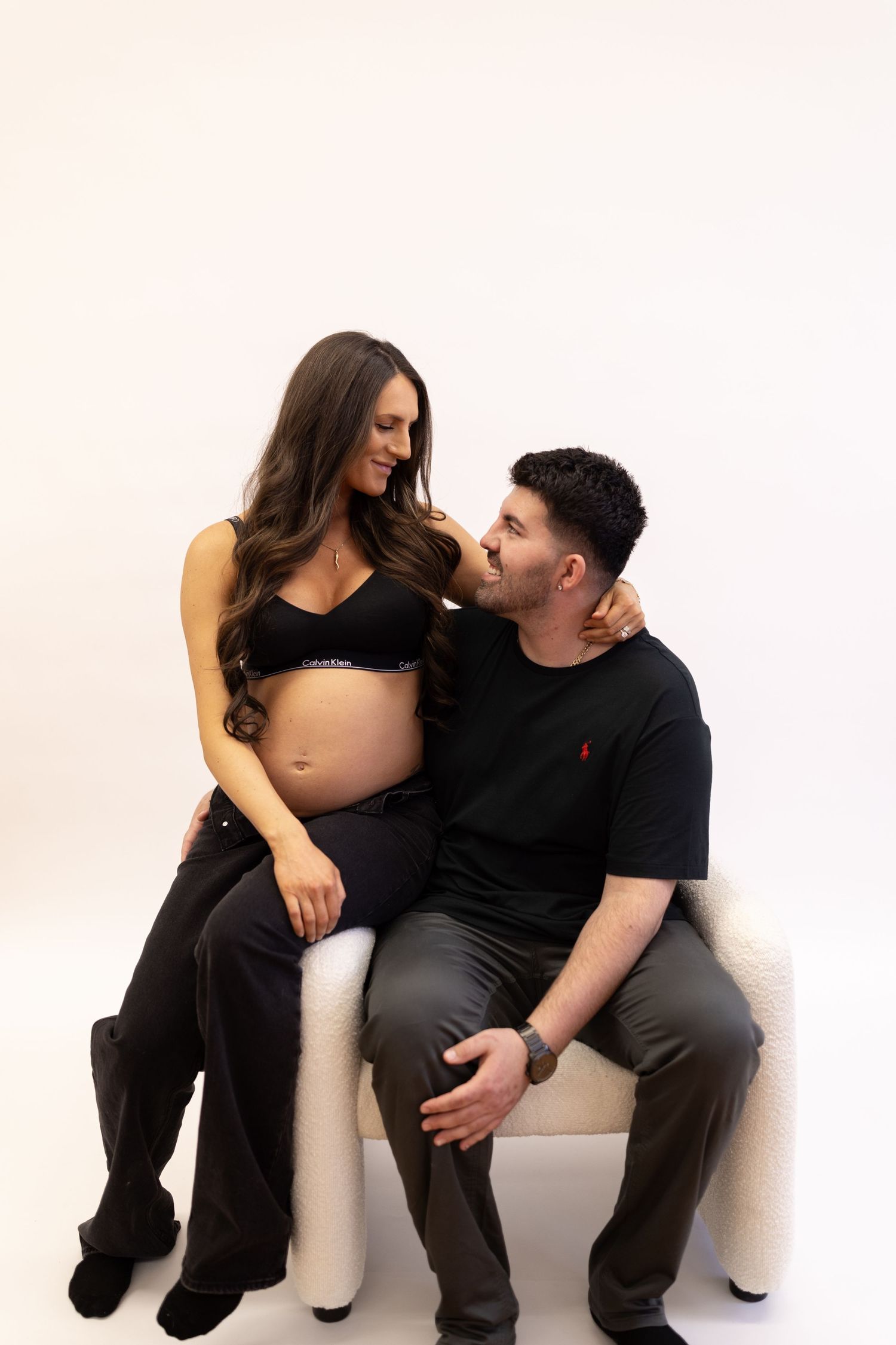 Pregnant woman in black Calvin Klein bralette sitting with partner on white chair, smiling lovingly.