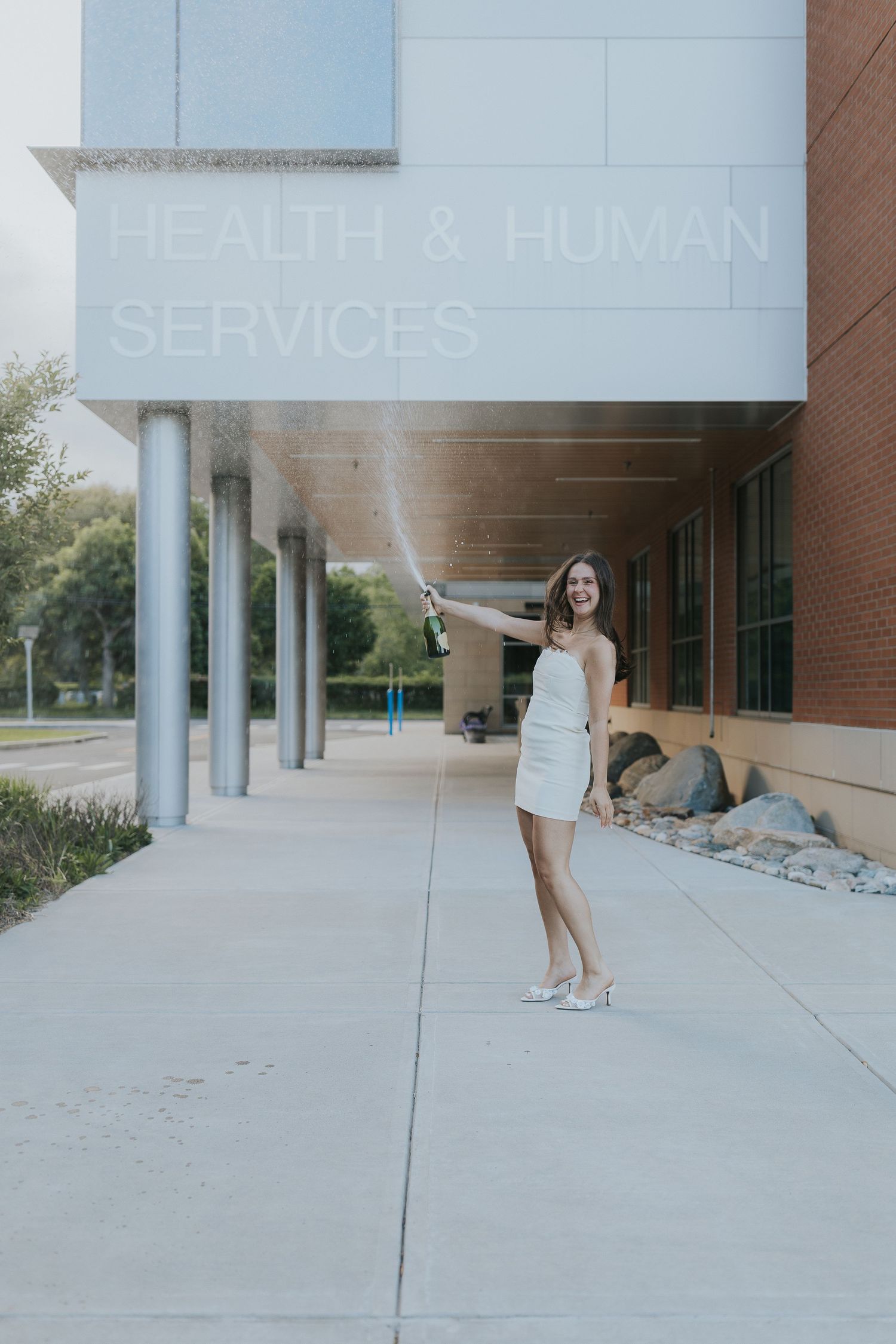 Woman in white dress celebrating graduation by popping champagne outside Health & Human Services building.