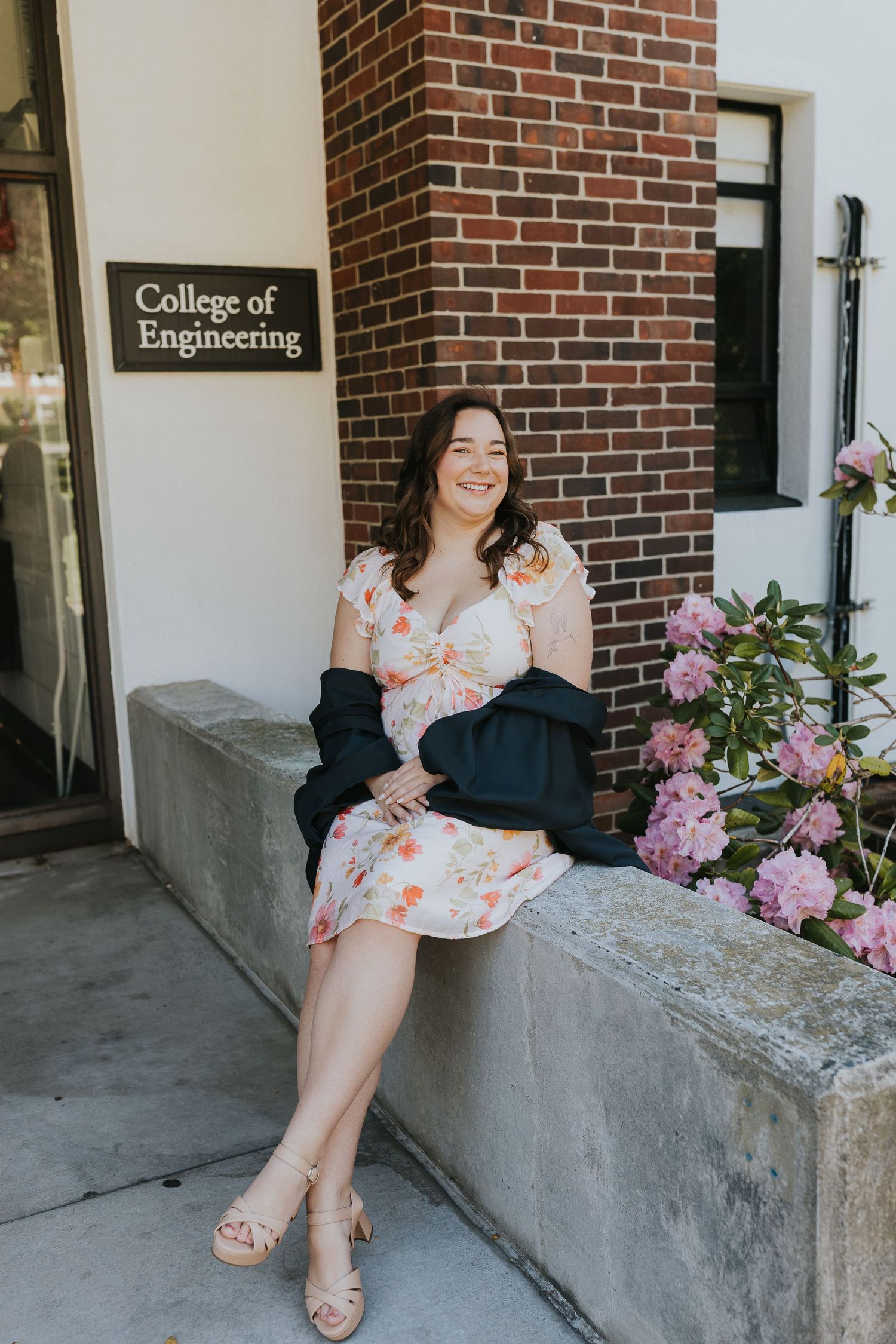 Smiling woman in floral dress seated outside College of Engineering building with pink flowers.