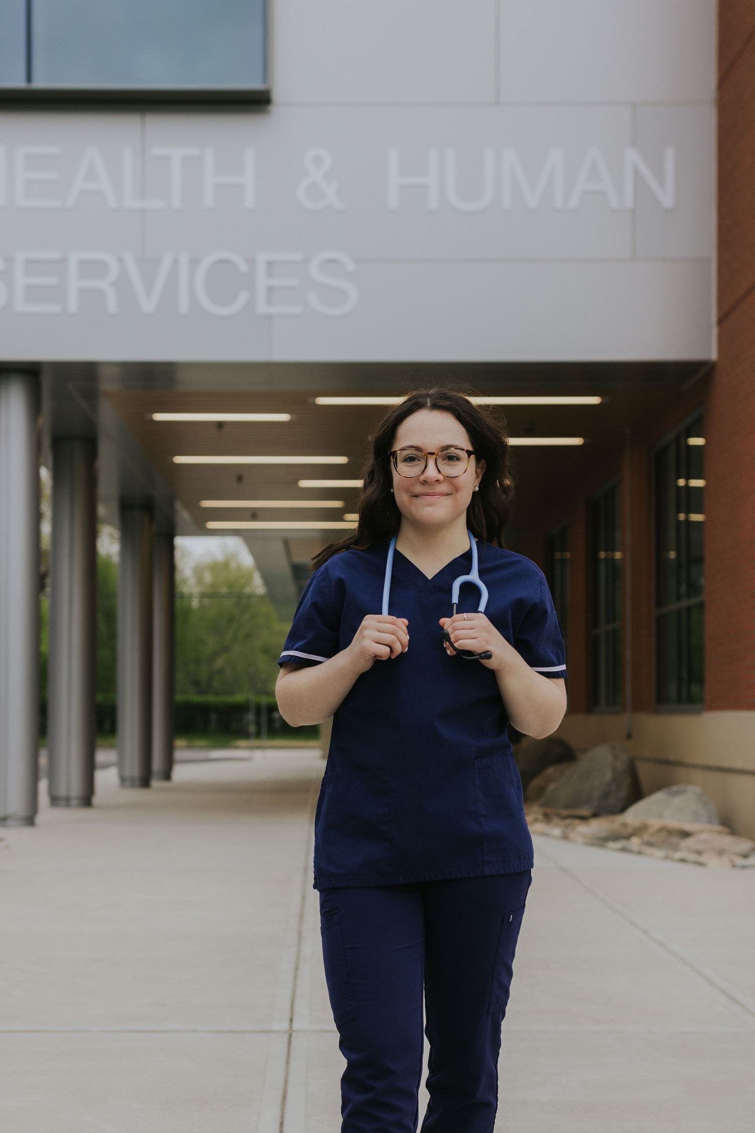 Female nursing student in navy blue scrubs with stethoscope stands outside Health and Human Services building.