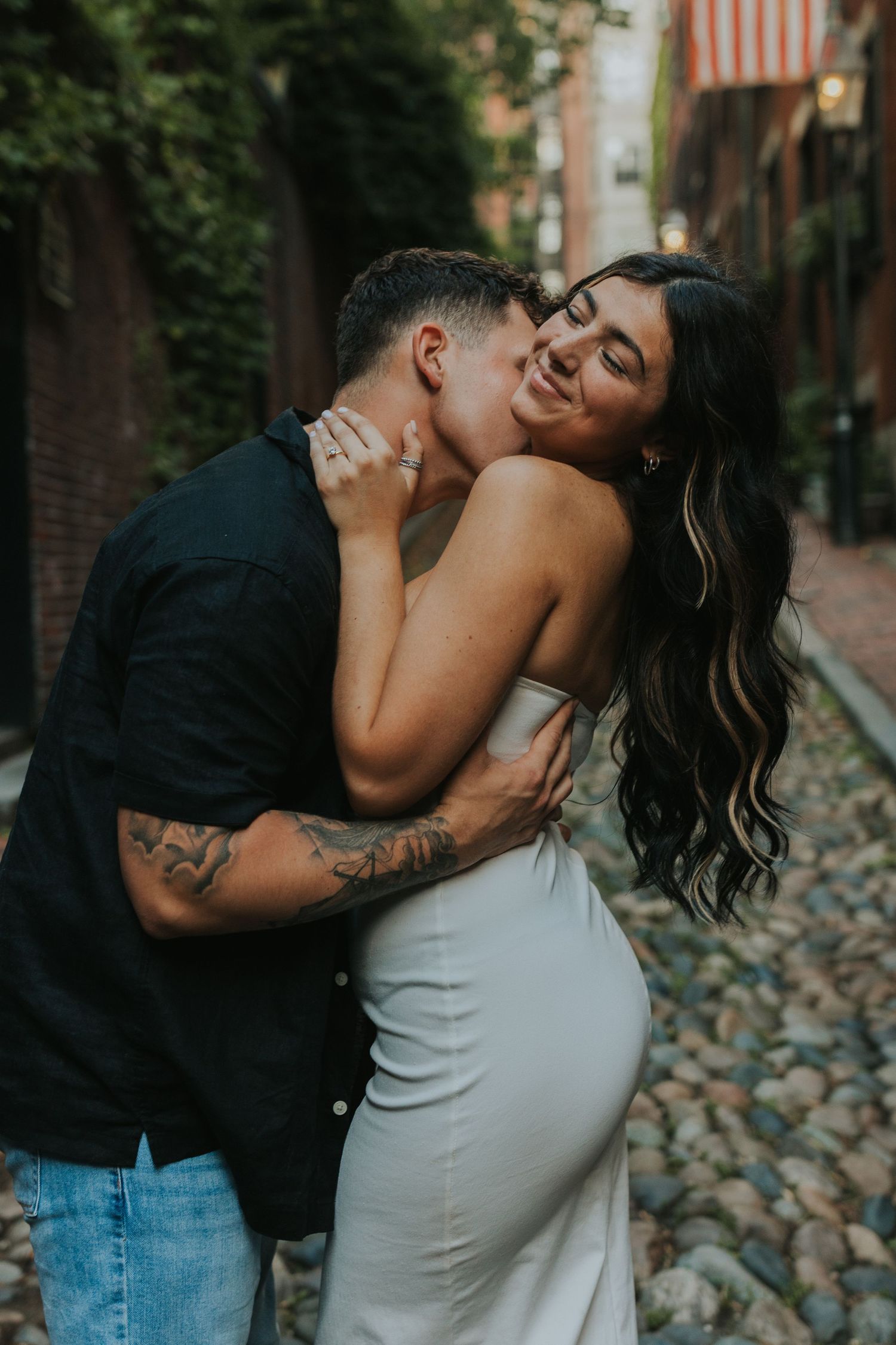 Man kissing woman on the neck as she smiles blissfully on a charming cobblestone street at dusk.