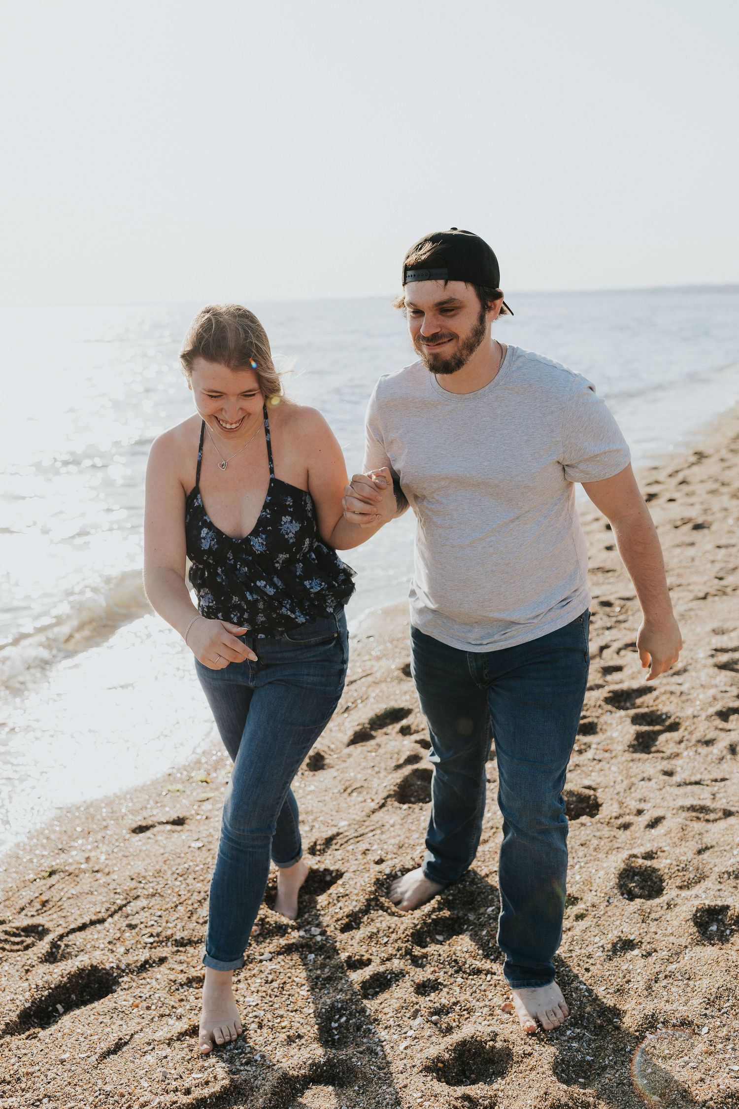 Smiling couple walks hand in hand along a sunny beach shoreline during engagement photos.