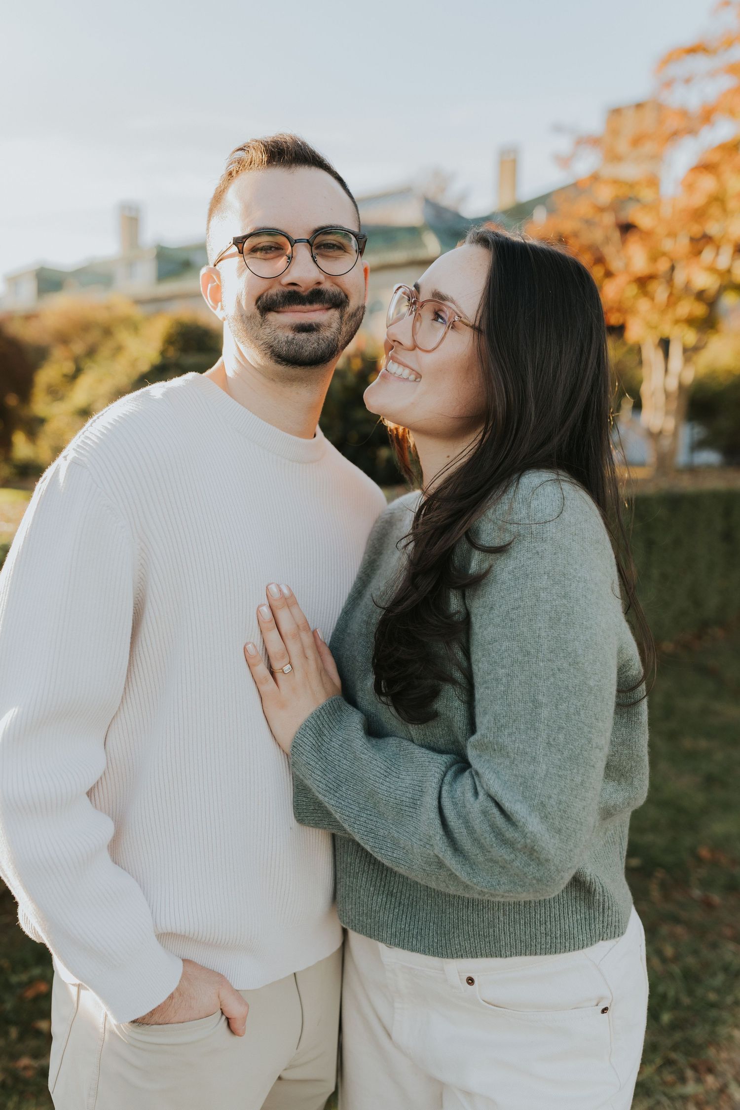 Smiling engaged couple outdoors in autumn, woman in gray sweater resting hand on man's chest showing engagement ring.