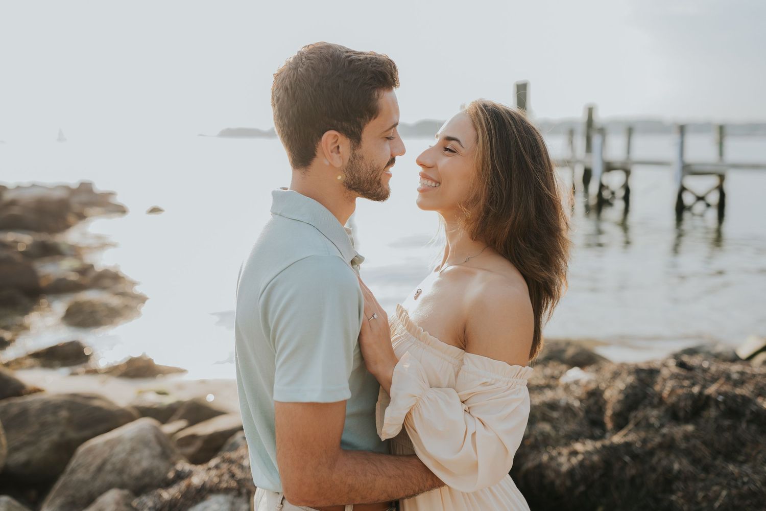 Couple sharing a tender moment on rocky shoreline, woman in off-shoulder dress embracing man in light blue polo.