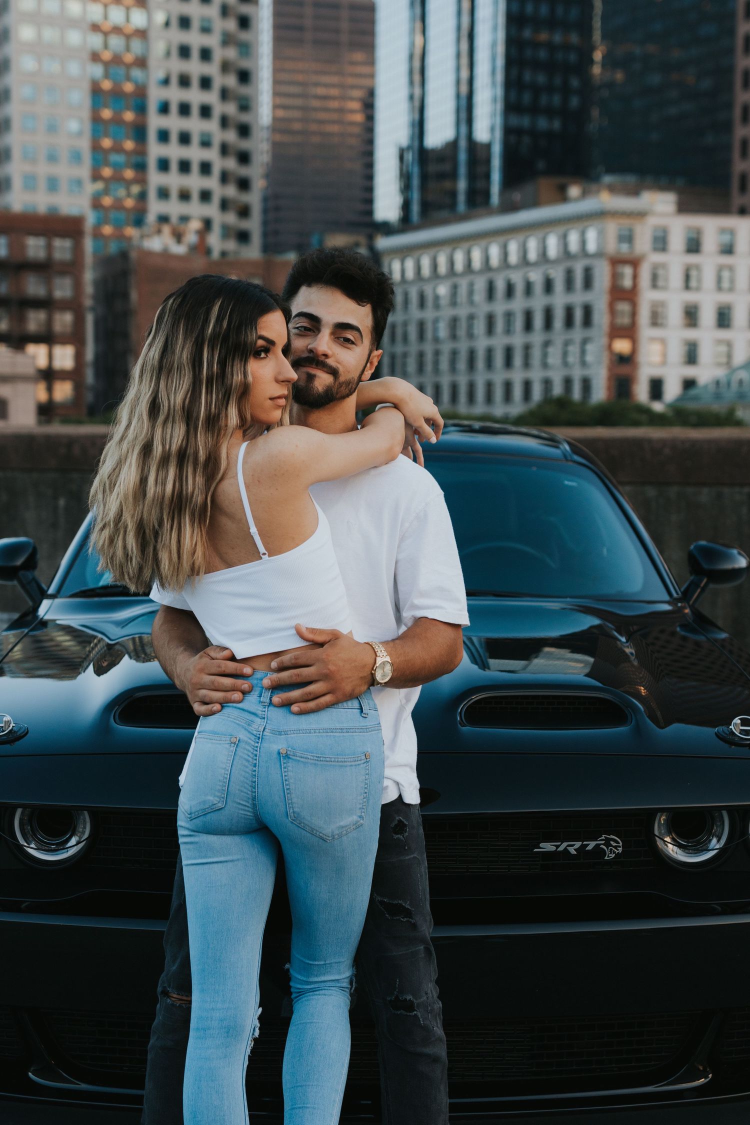 Stylish couple posing together in front of a black Dodge SRT muscle car with city skyline backdrop.