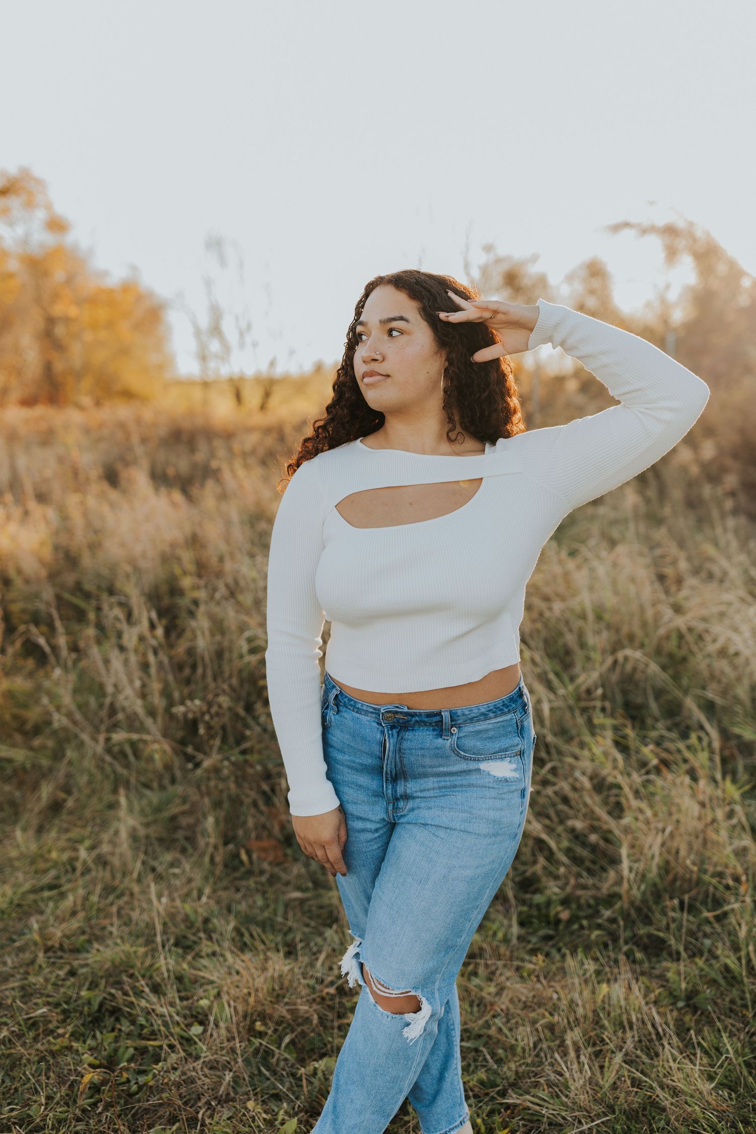 Young woman in white cutout crop top and ripped jeans posing in autumn field at sunset.