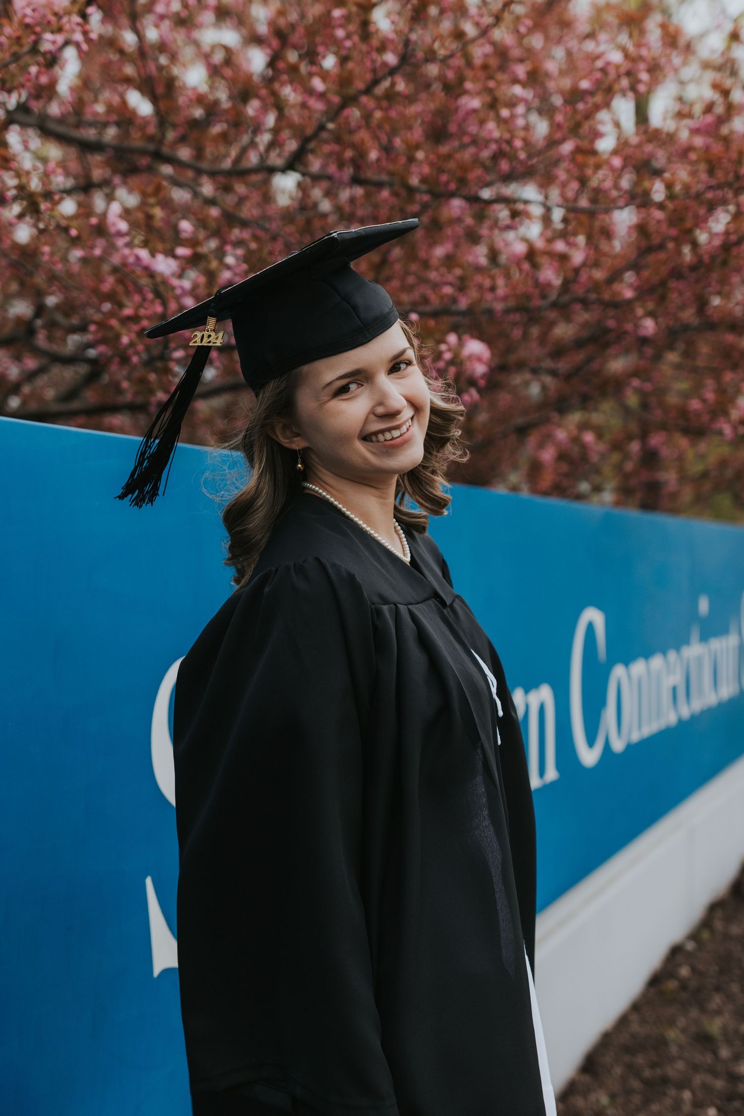 Smiling female graduate in cap and gown poses beside Southern Connecticut blue banner with cherry blossoms.